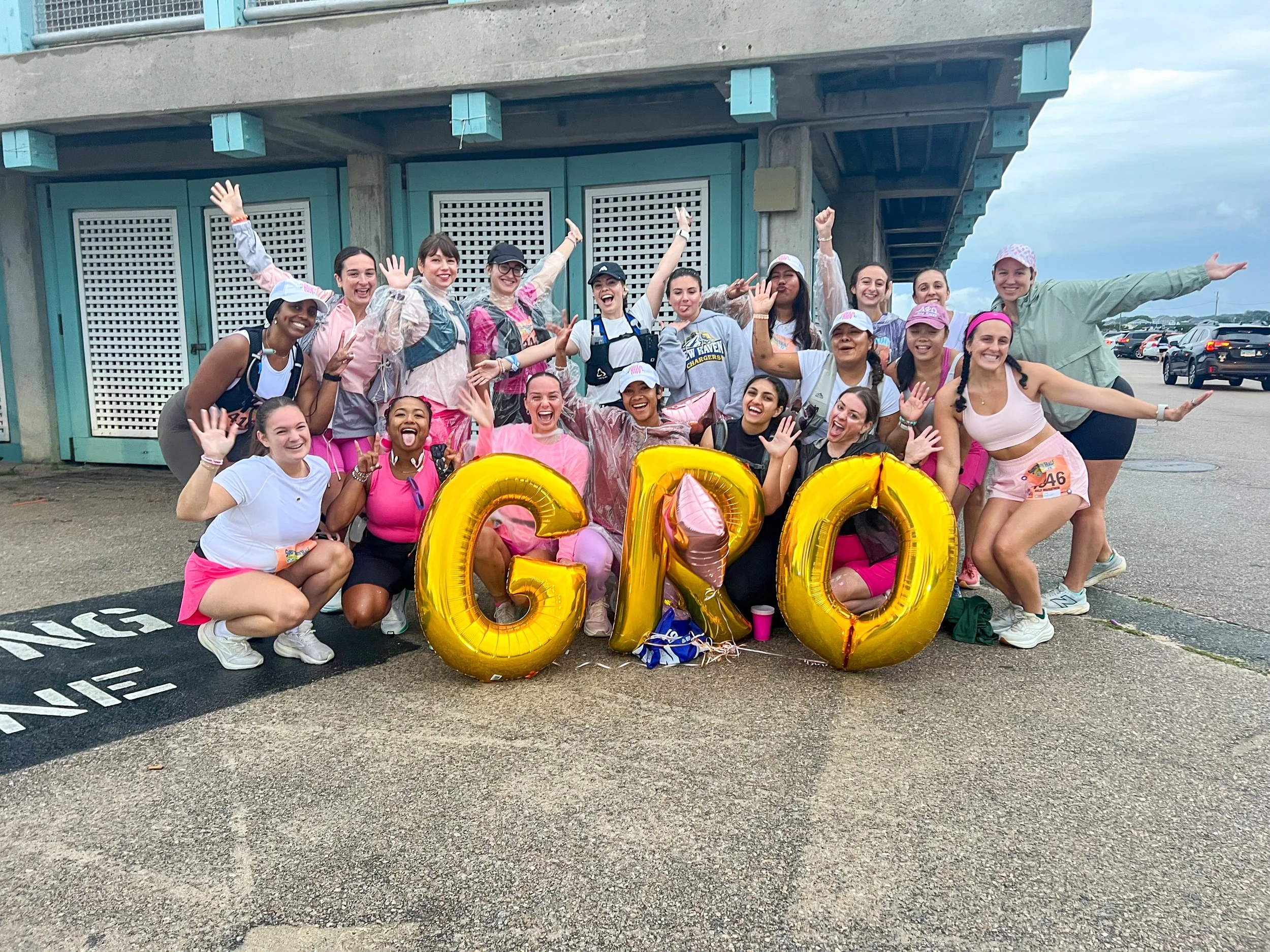 A group of women, dressed in pink and athletic wear, celebrating and posing with large gold balloon letters spelling GNO outside near a parking lot and a building.