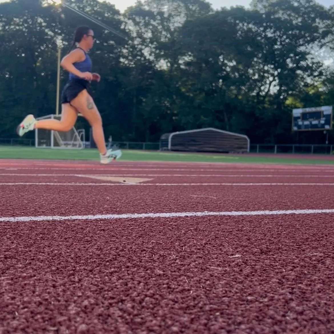 A woman running on a red track field during daytime with trees and a scoreboard in the background.