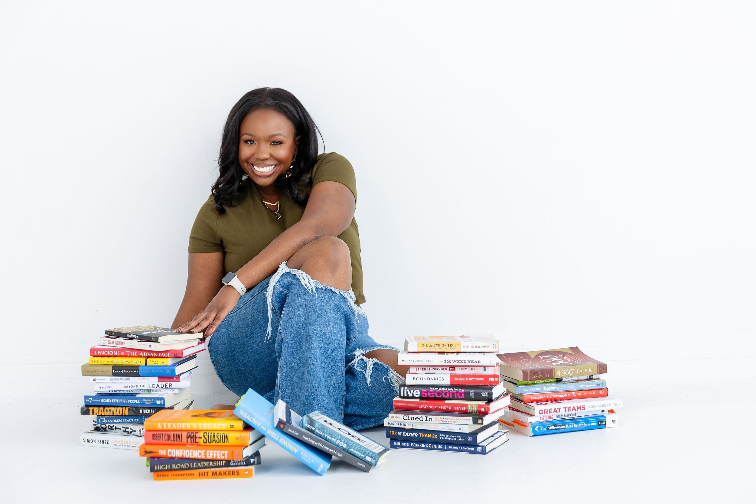 A smiling woman with long black hair, wearing a green t-shirt, ripped jeans, and a smartwatch, sitting cross-legged surrounded by stacks of books on a white background.