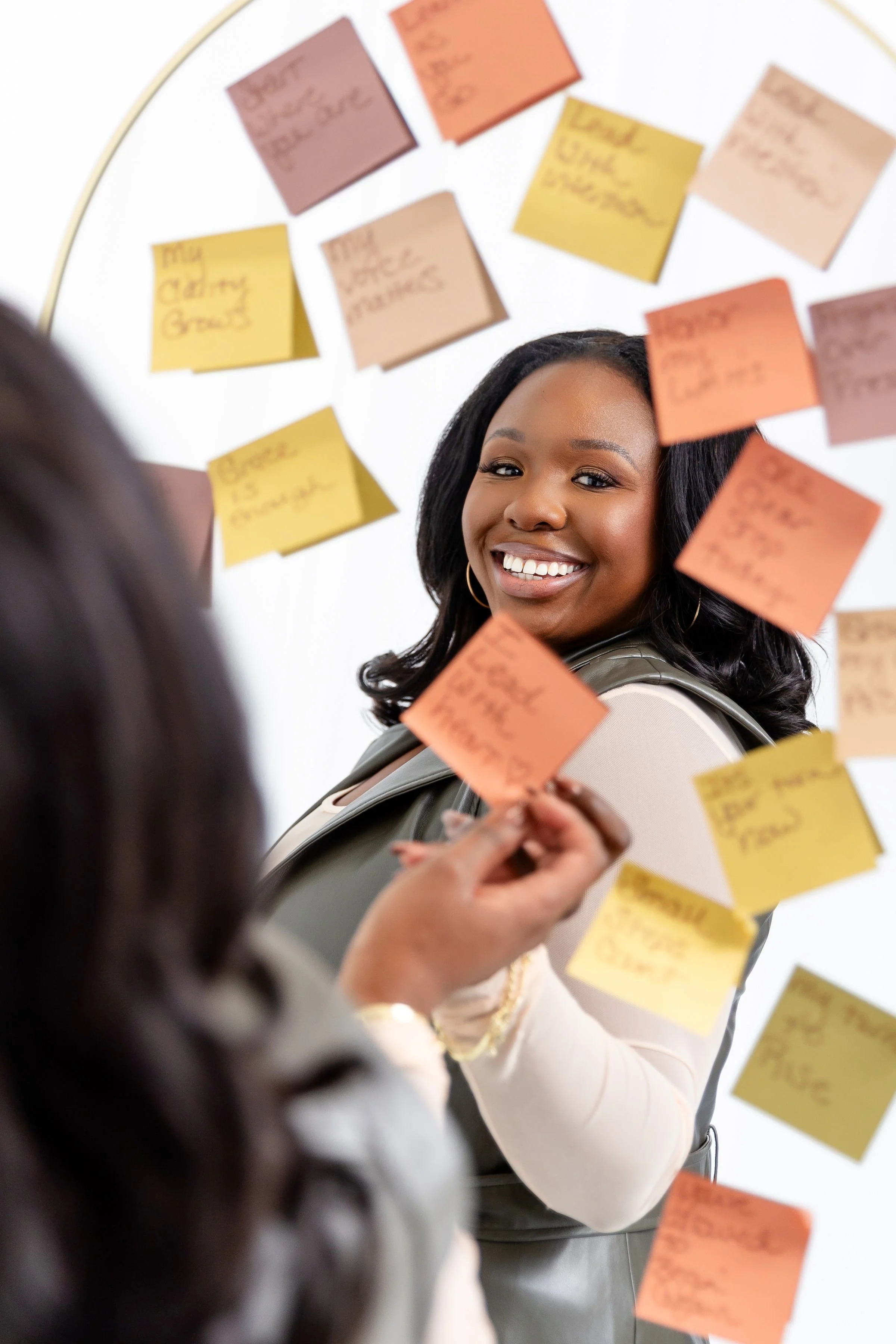 Two women in a business setting are discussing ideas written on colorful sticky notes on a wall, with one woman smiling at the camera.