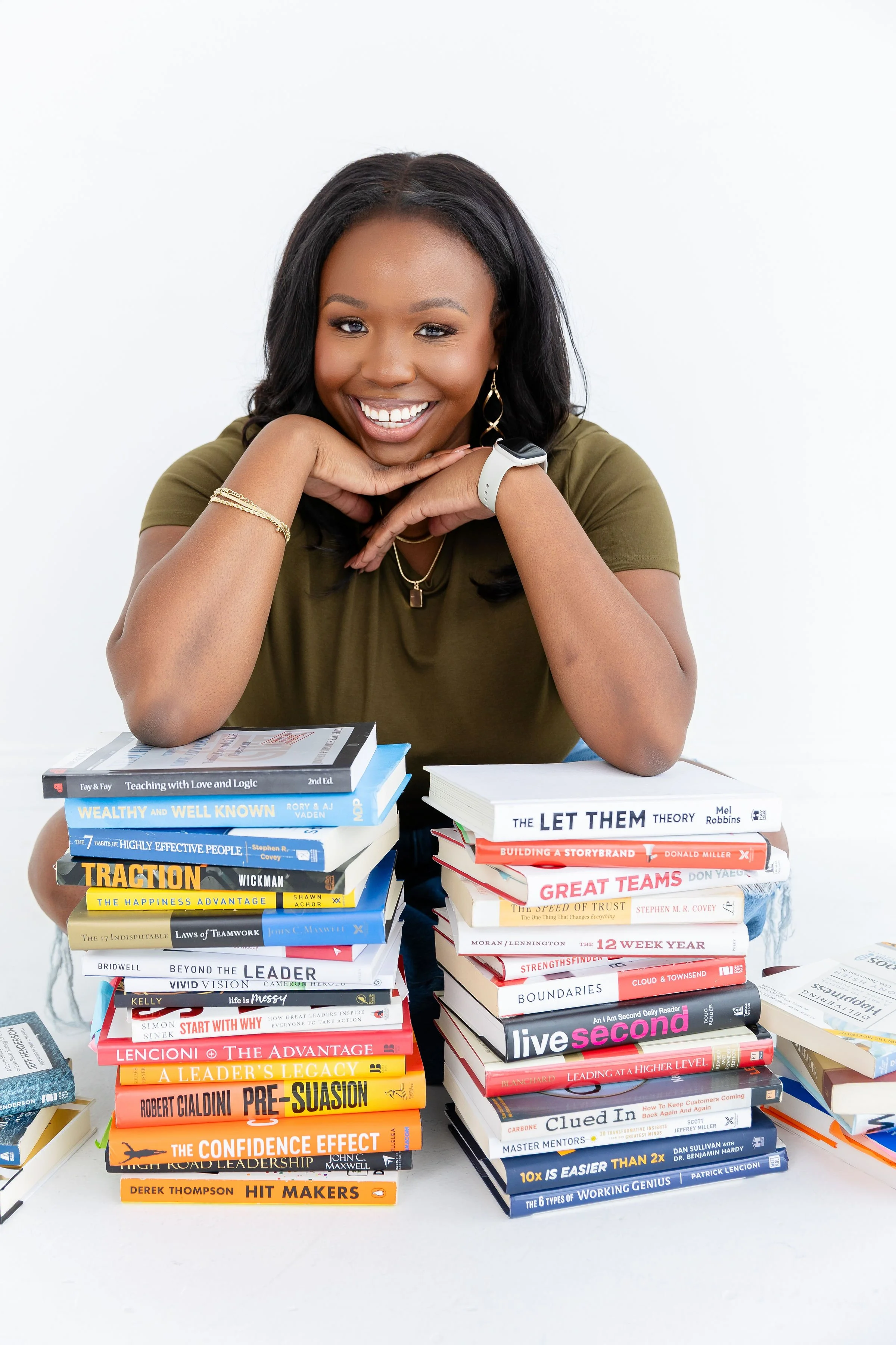 A woman smiling and sitting at a table with multiple stacks of books in front of her.