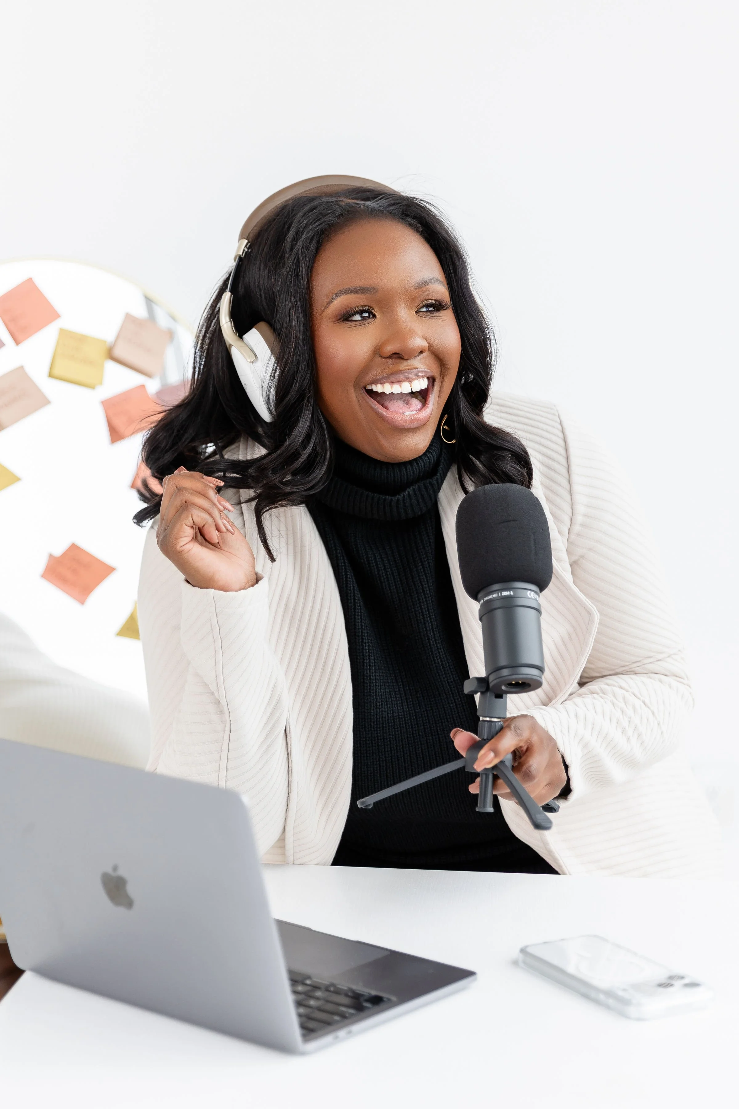 A young woman with dark hair, wearing headphones, holding a microphone, smiling and speaking in front of a laptop and smartphone on a white desk.