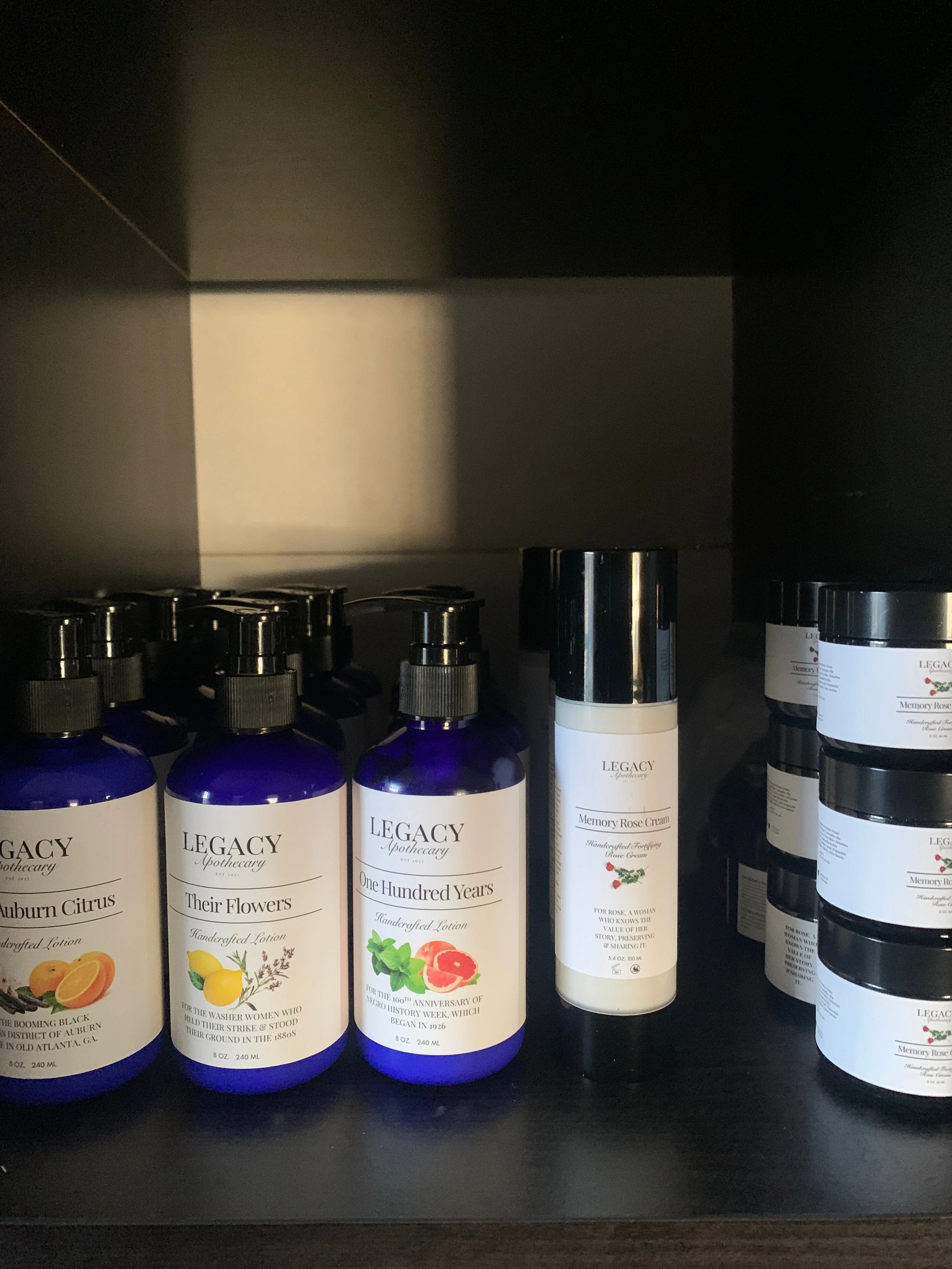 Shelf with blue bottles labeled 'Legacy Apothecary' containing various handcrafted lotions and creams, including Auburn Citrus, Their Flowers, One Hundred Years, and Memory Rose Cream.