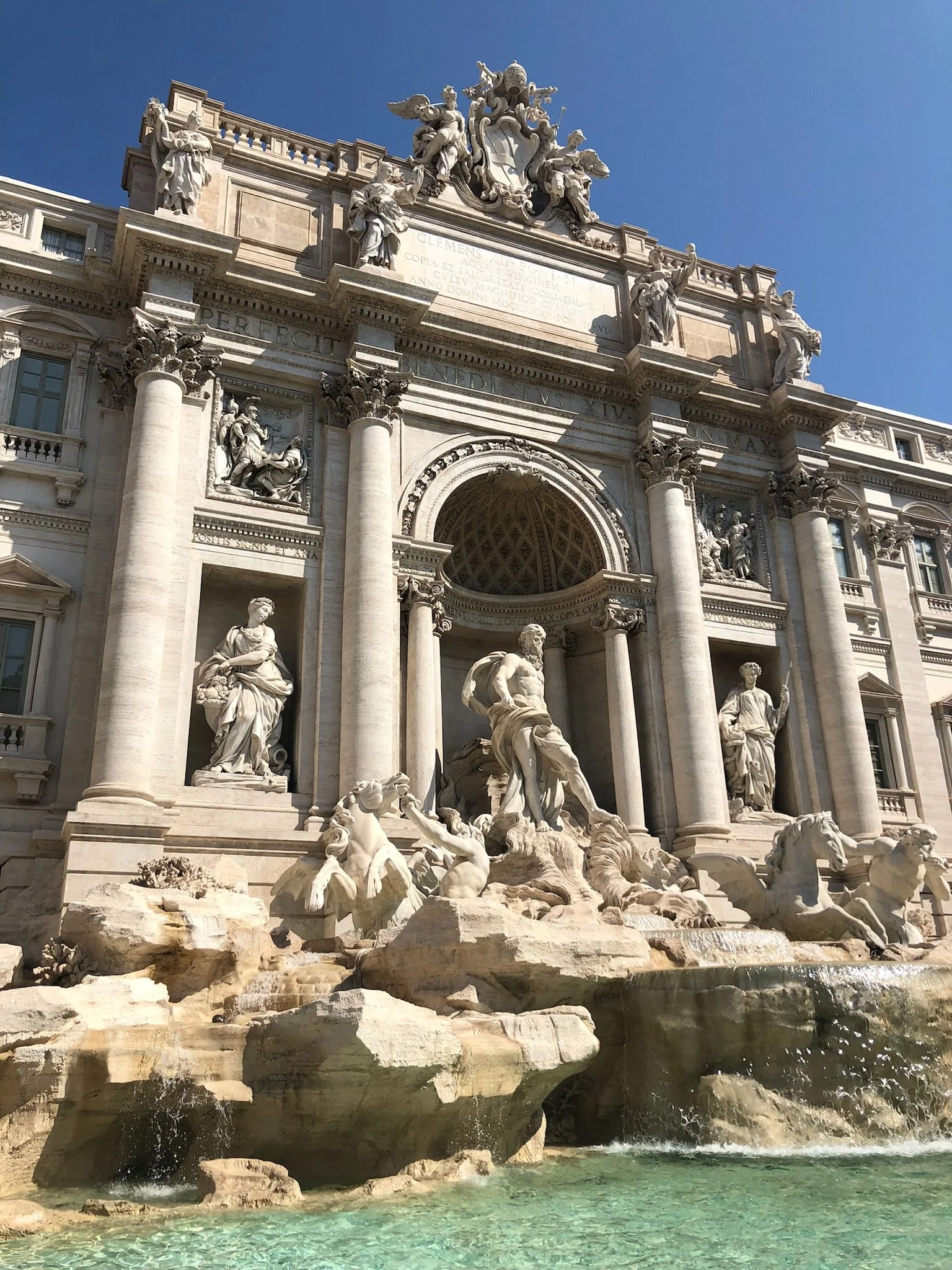 The Trevi Fountain in Rome, Italy, showcasing elaborate sculptures and cascading water at the base. The architecture features large columns and detailed carvings under a clear blue sky.