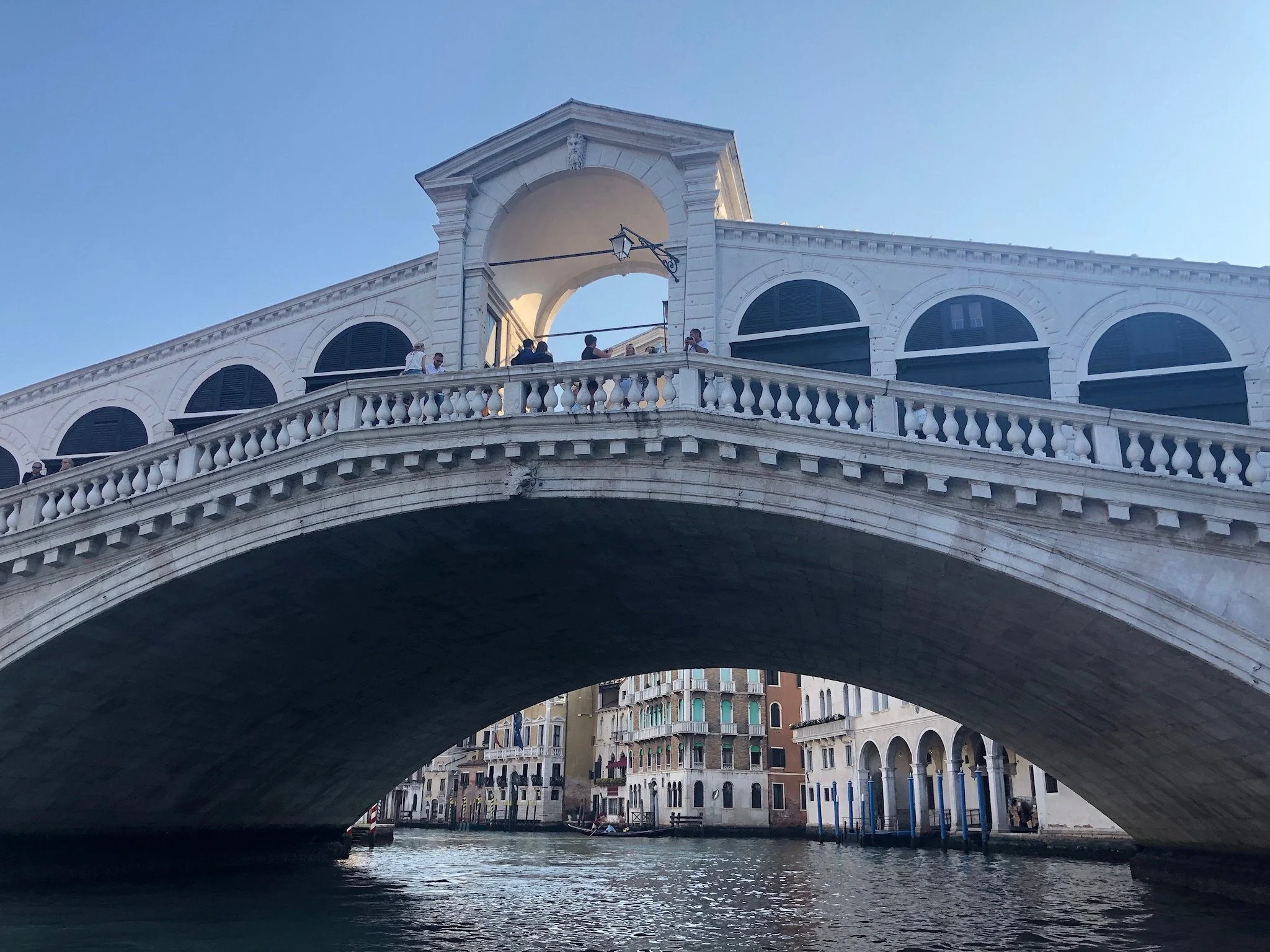 View of a historic bridge over a canal in Venice, Italy, with buildings and people on the bridge, clear blue sky.