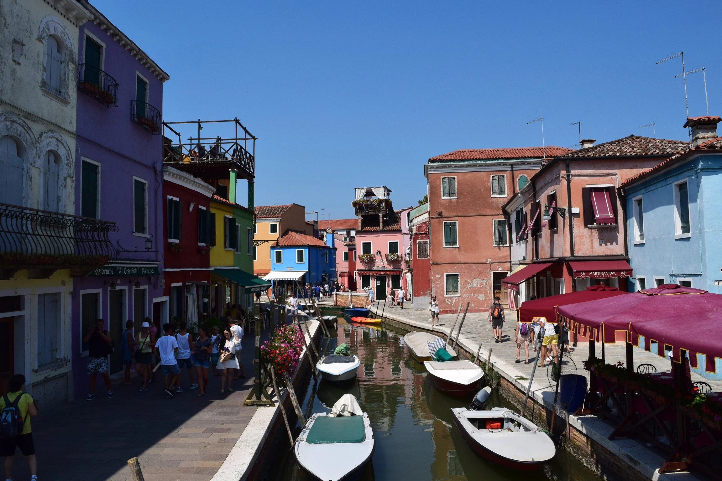 Colorful buildings along a canal with boats in a tourist area, under a clear blue sky.