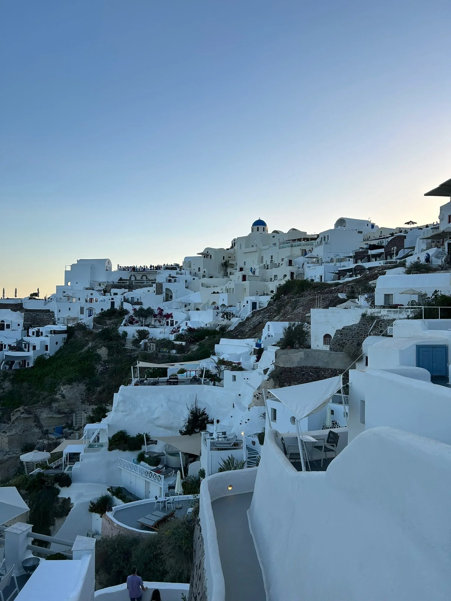 Sunset view of whitewashed buildings on a hillside in Santorini, Greece, with a clear blue sky above.