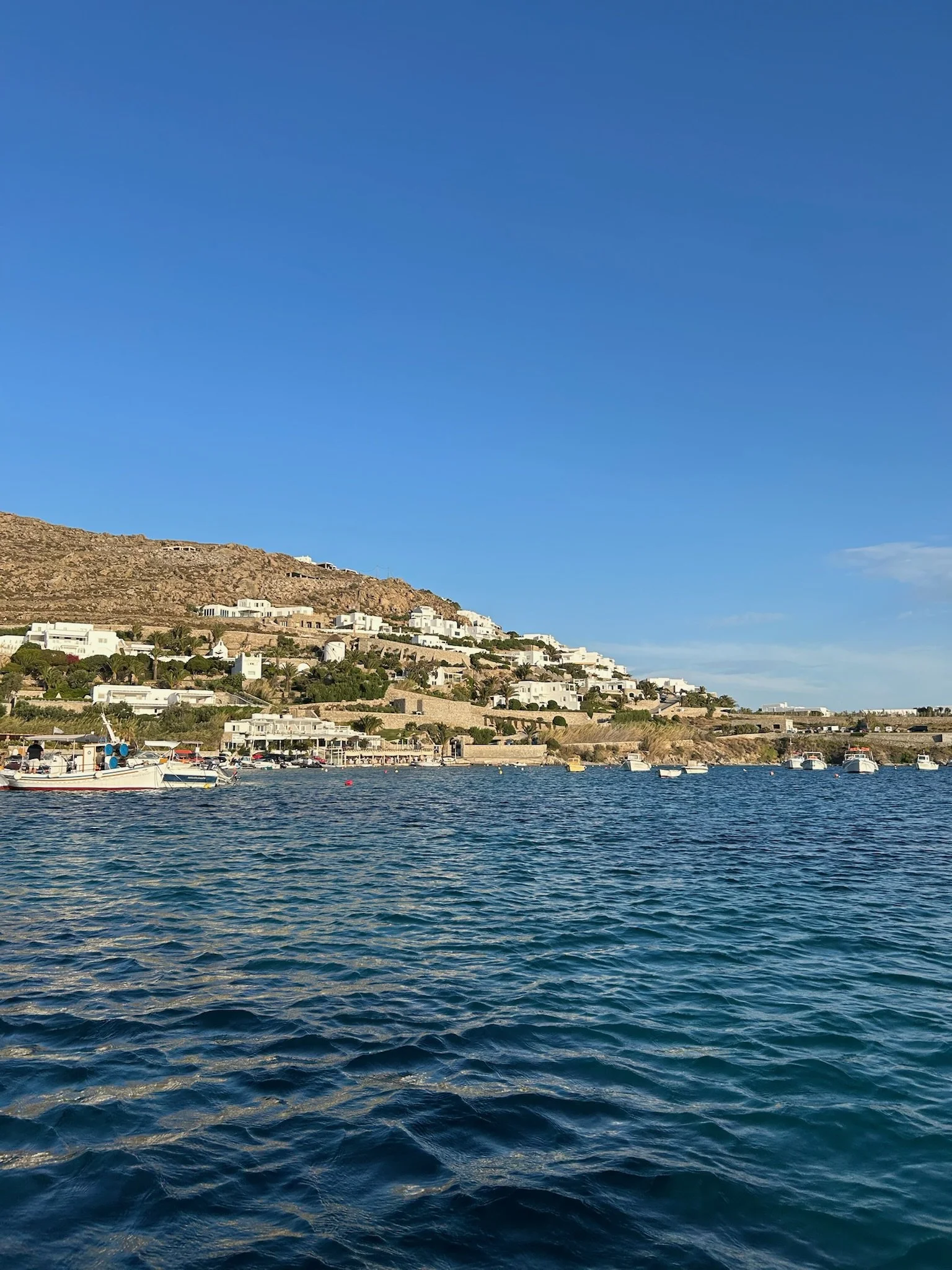 A coastal scene with calm blue water, several boats docked near the shoreline, and white buildings on a hillside under a clear blue sky.