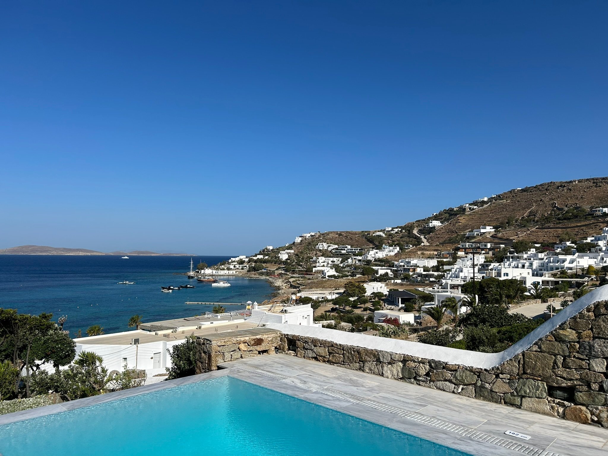 Scenic view of a coastal village with white buildings on a hillside, blue sea with boats, and a clear sky, seen from a swimming pool in the foreground.