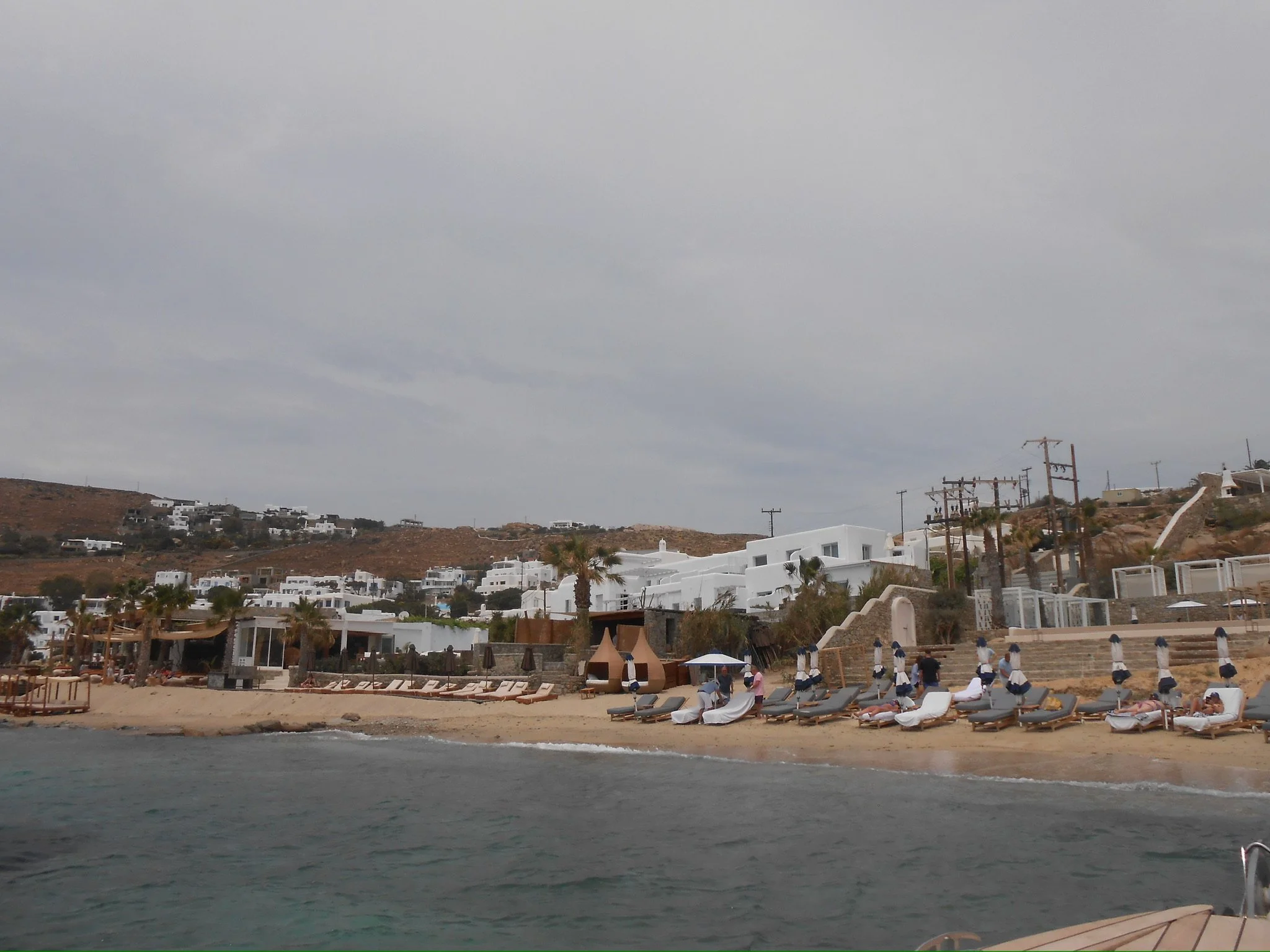 A beachside resort with lounge chairs, umbrellas, white buildings on a hillside, and power lines on a cloudy day.