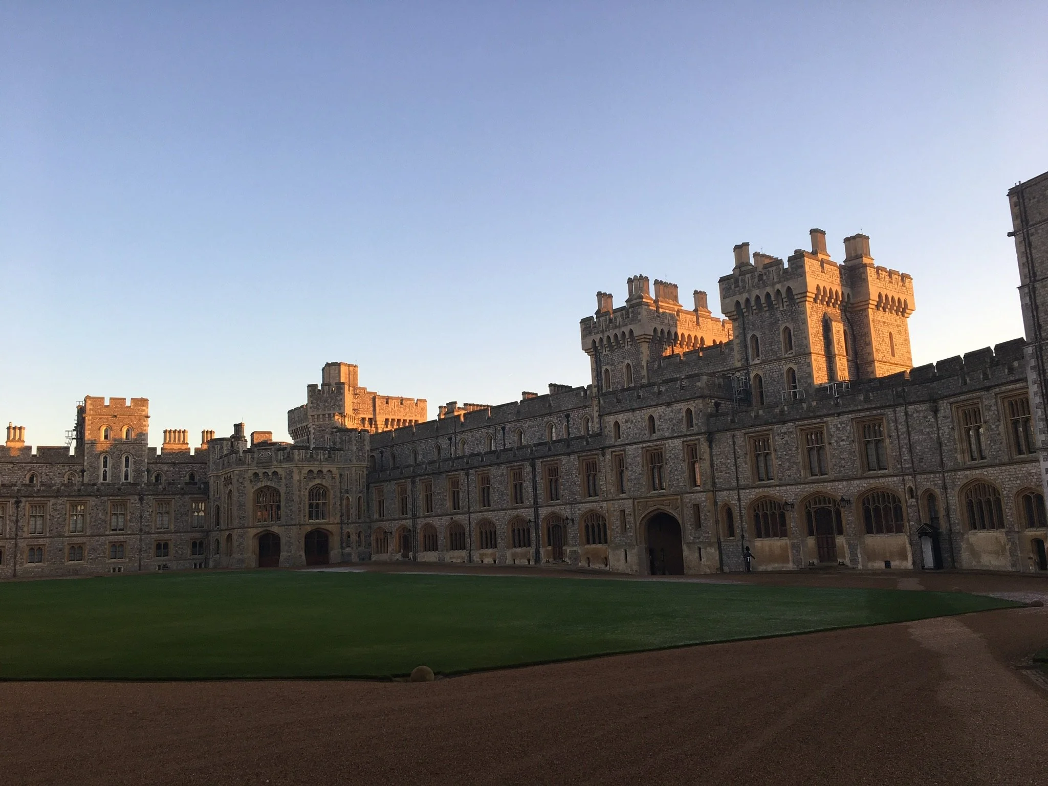 A large castle with multiple towers and crenellated walls, built with stone, set against a clear sky, with a grass lawn and a dirt path in the foreground.