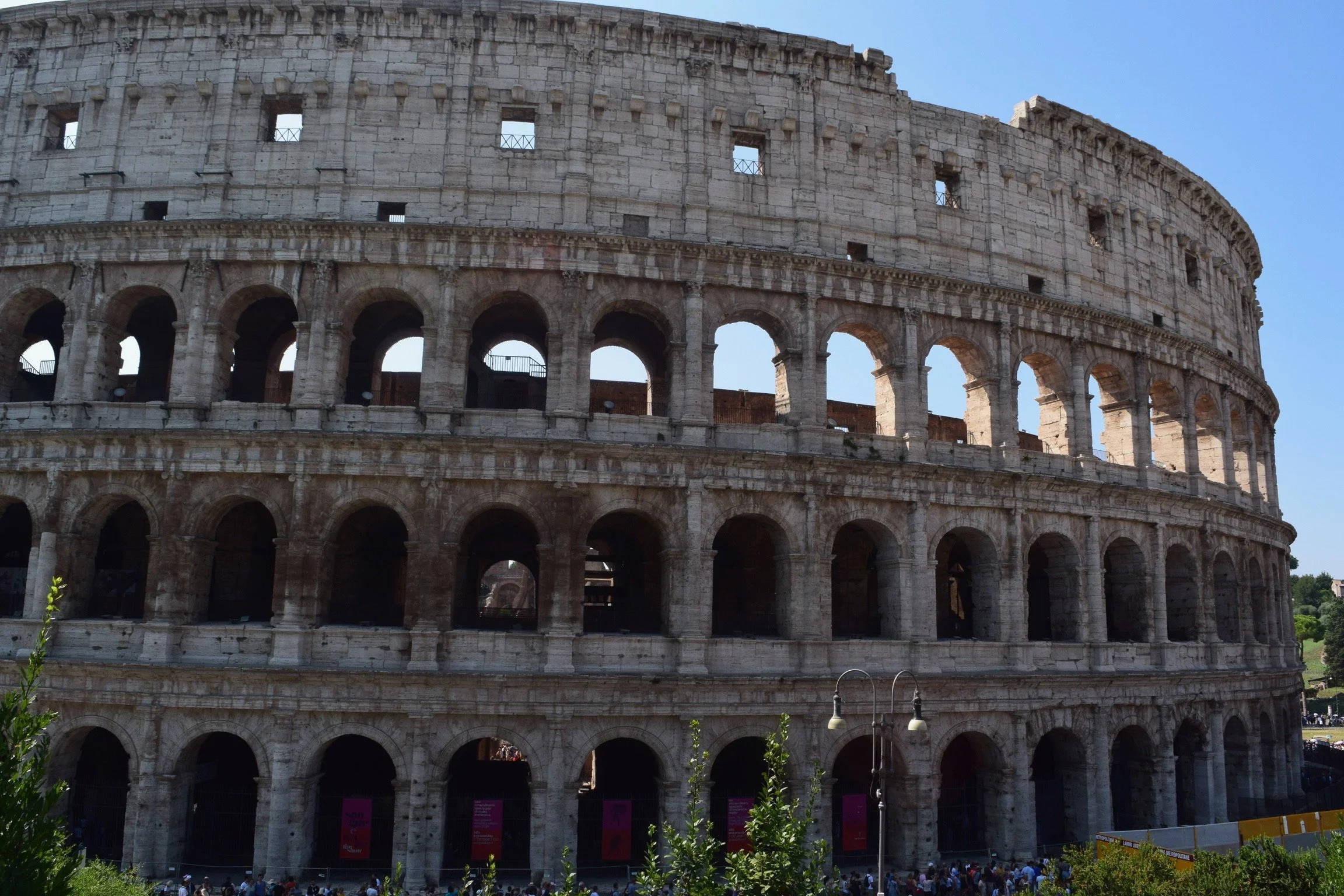 Exterior view of the Colosseum in Rome, Italy, showing its stone structure and multiple arched openings, with visitors gathered at its base.