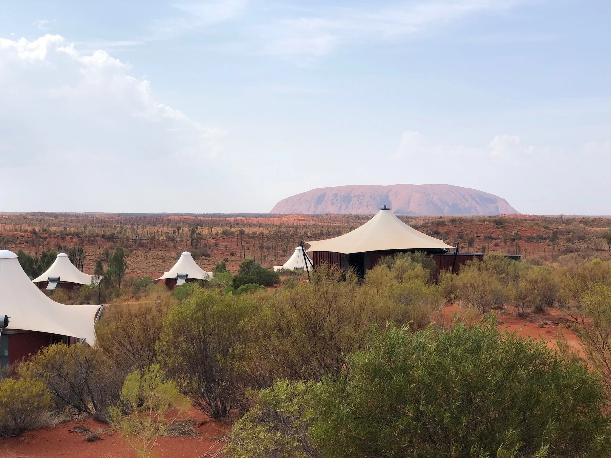 Desert landscape with dome-shaped tents and sparse vegetation, with Uluru rock formation in the background under a partly cloudy sky.