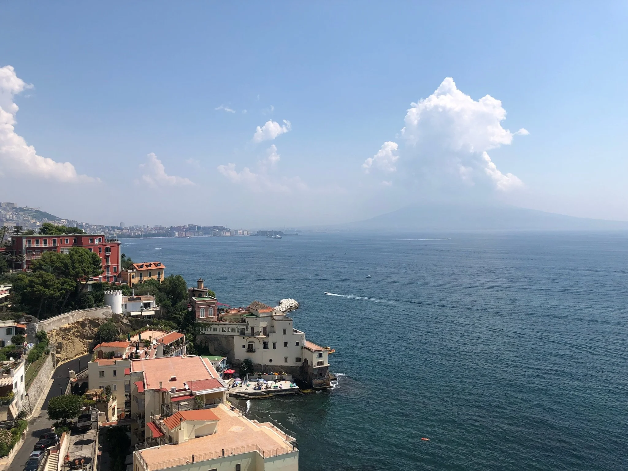 Coastal cityscape with colorful buildings on cliffs overlooking the ocean, Mount Vesuvius in the distance, and boats on the water.
