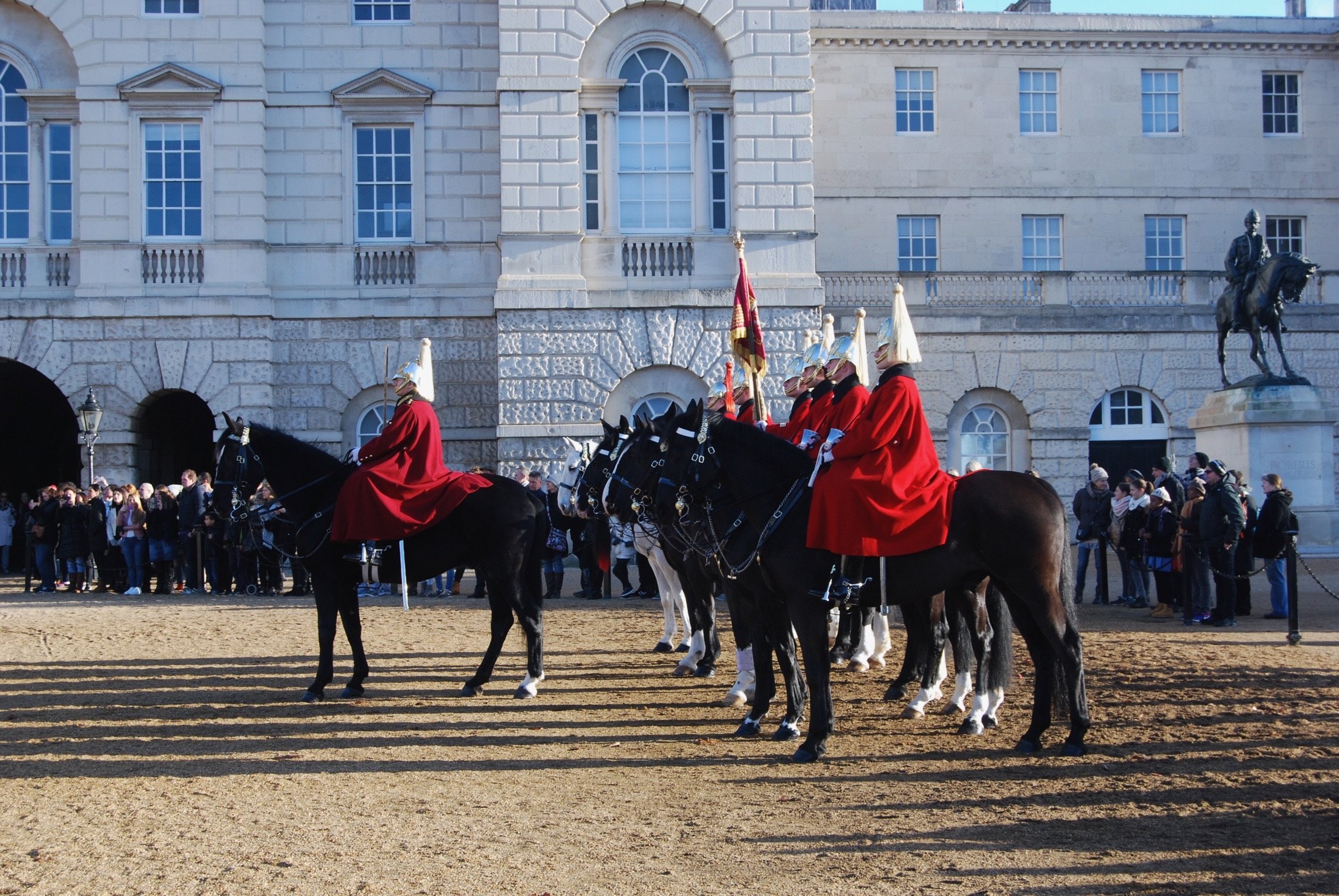 Mounted guards in red cloaks participating in a ceremonial event outside a historic stone building, with spectators watching.