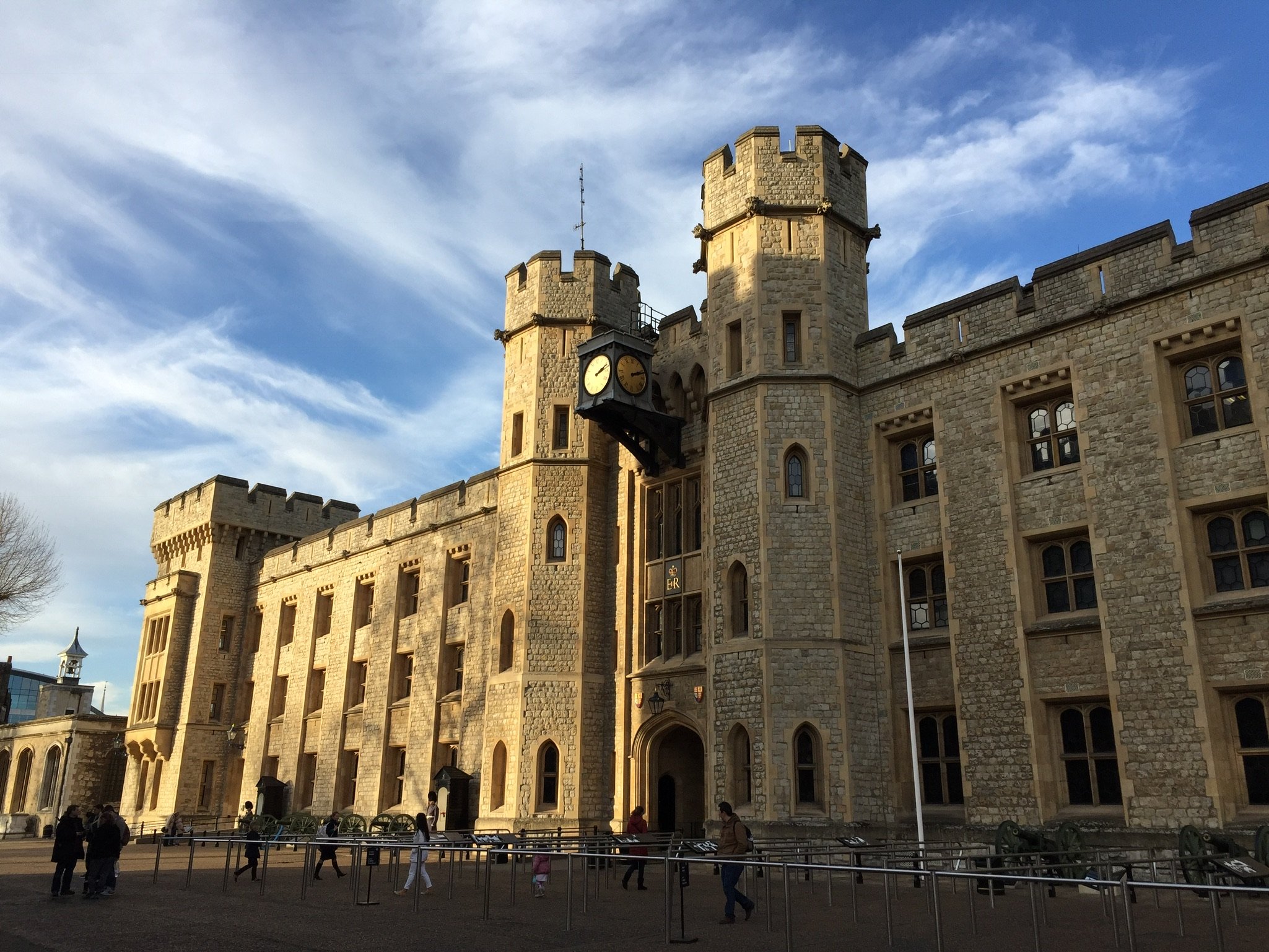 A historic stone castle with towers and a clock, under a partly cloudy sky, with people walking in front.