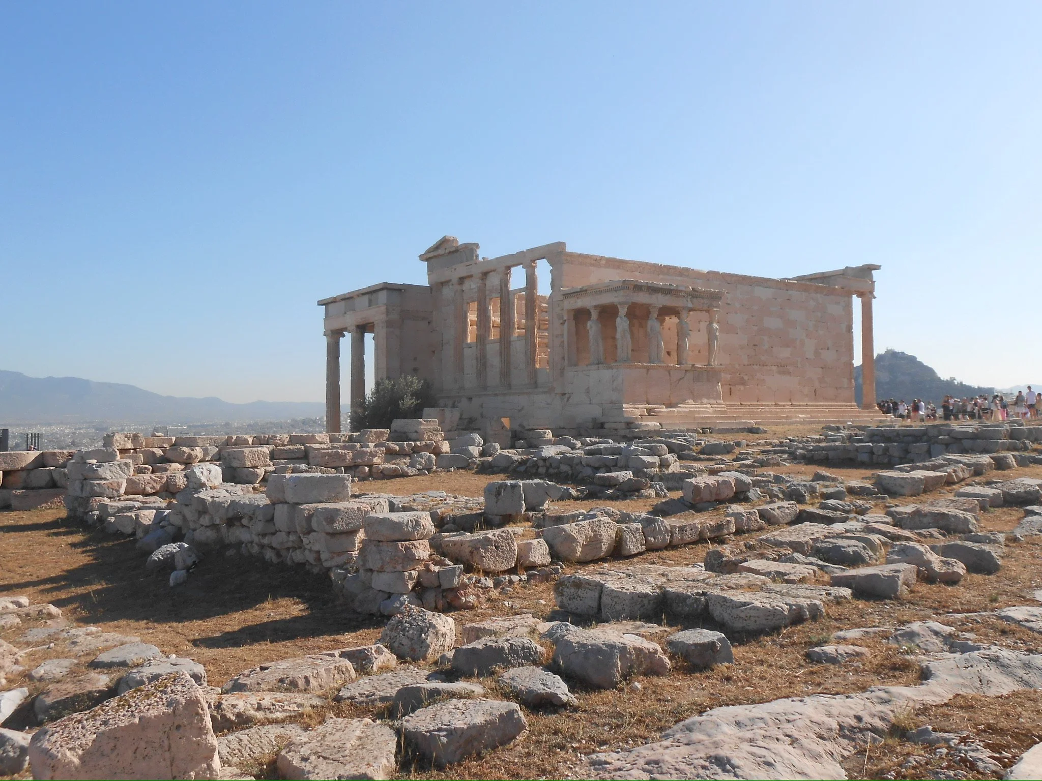 Ancient Greek ruins with columns and collapsed stones on arid ground, with a mountain and cityscape in the background under a clear blue sky.