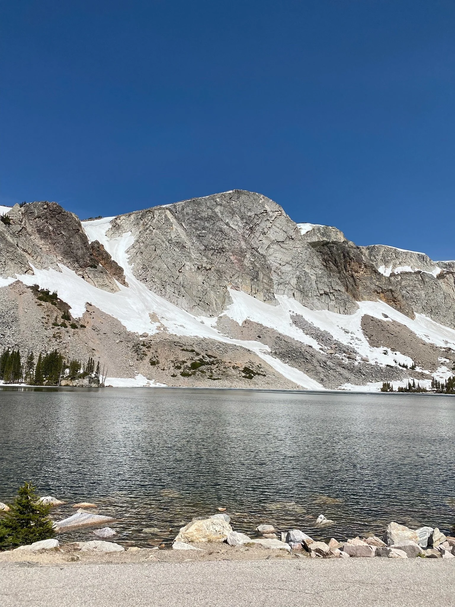 A view of a mountain with patches of snow, a lake in the foreground, and a clear blue sky.