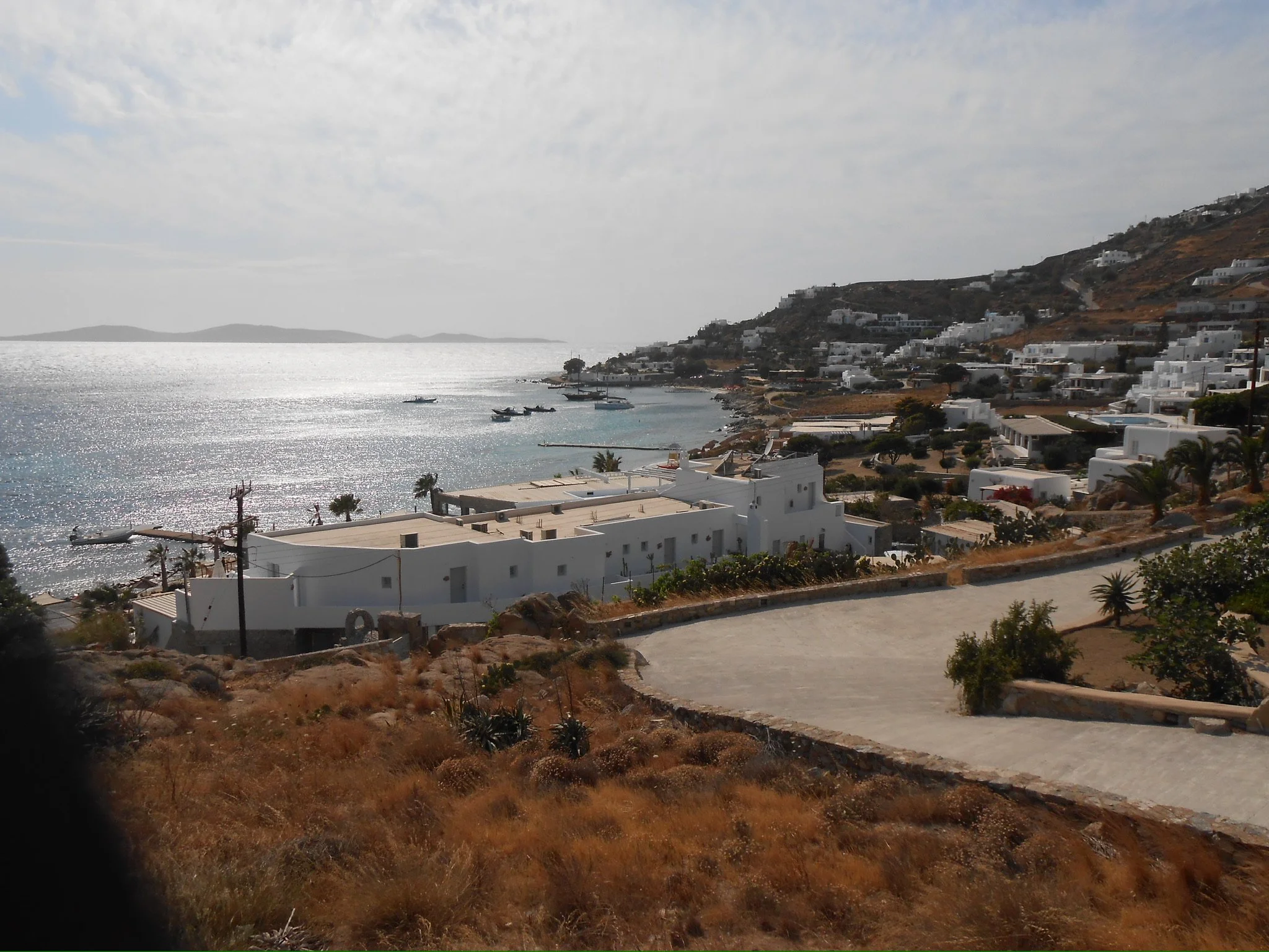 Coastal view of white buildings on a hillside overlooking the ocean, with boats in the water and a partly cloudy sky.