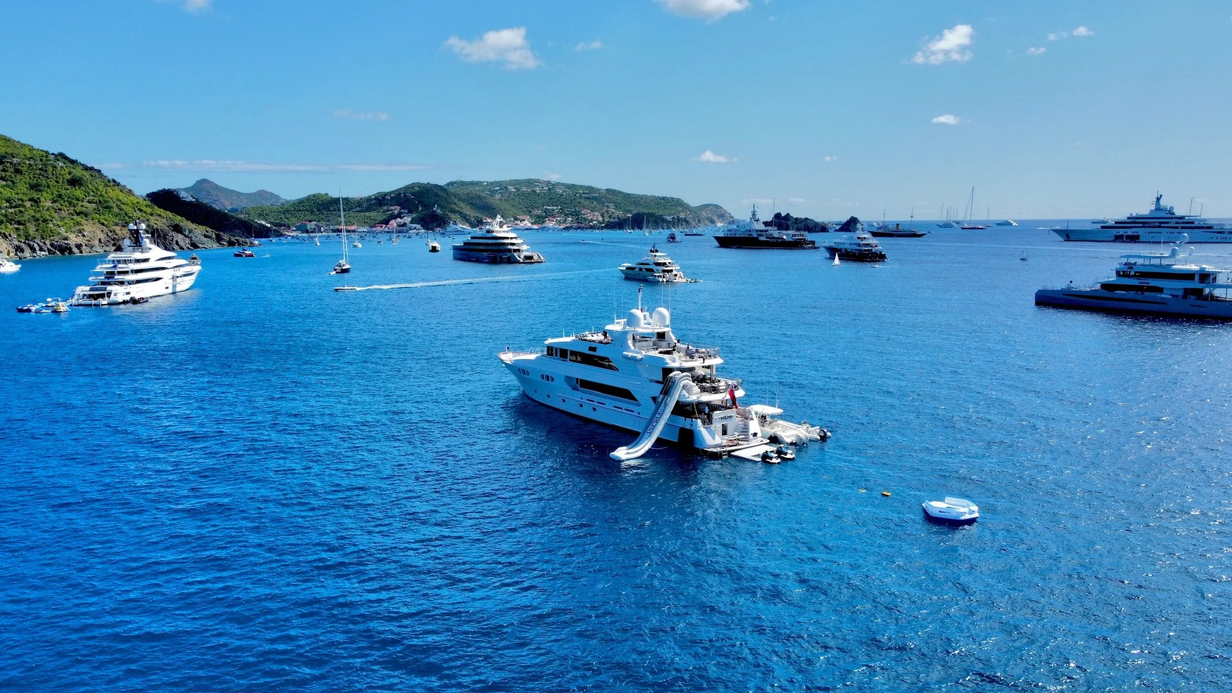A harbor with several large yachts and boats anchored on calm blue water, with a hilly landscape and small islands in the background under a partly cloudy sky.