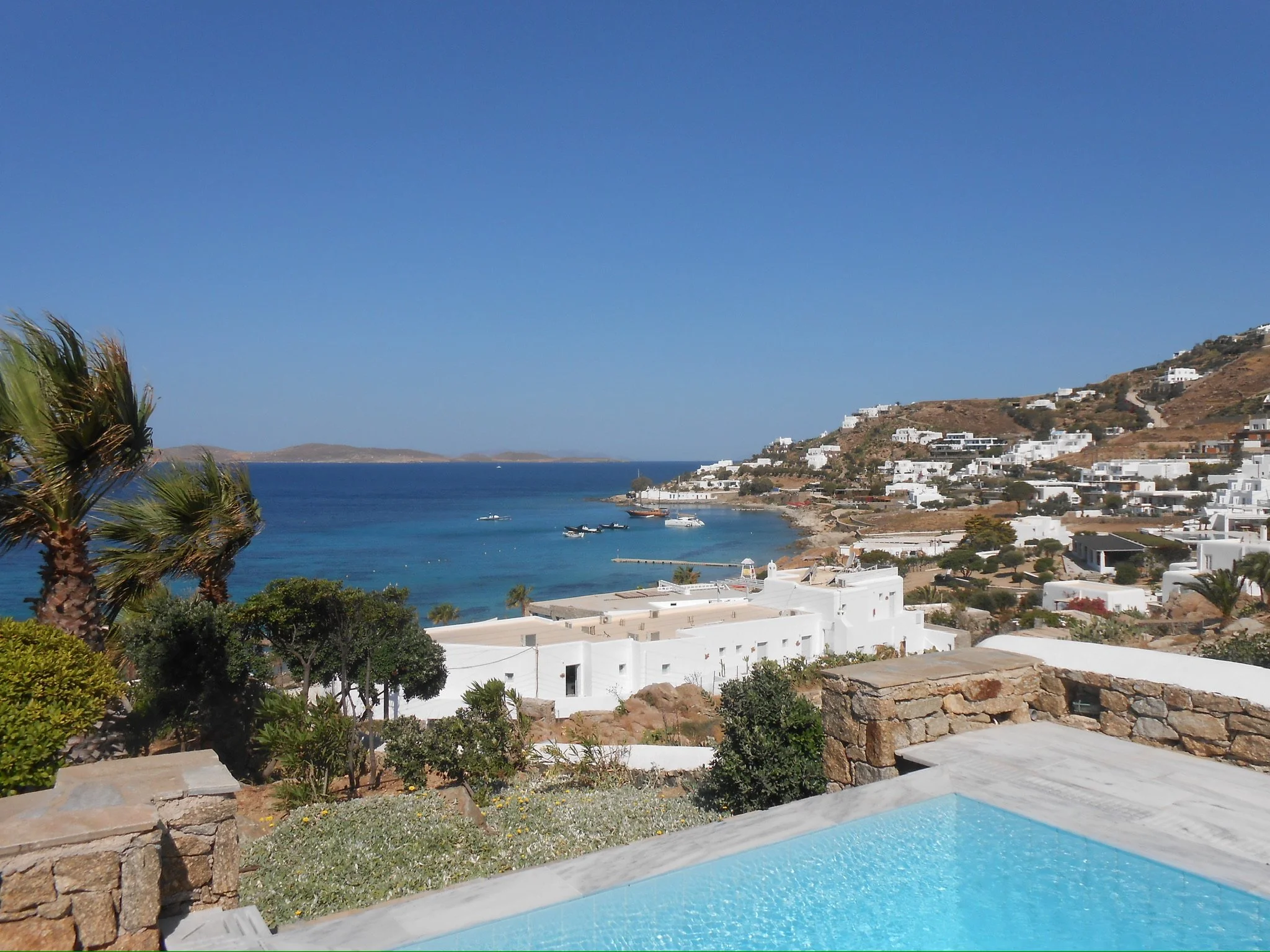 Scenic view of a coastal village with white buildings on a hillside overlooking a bay with boats, clear blue water, and a small pool in the foreground under a bright blue sky.