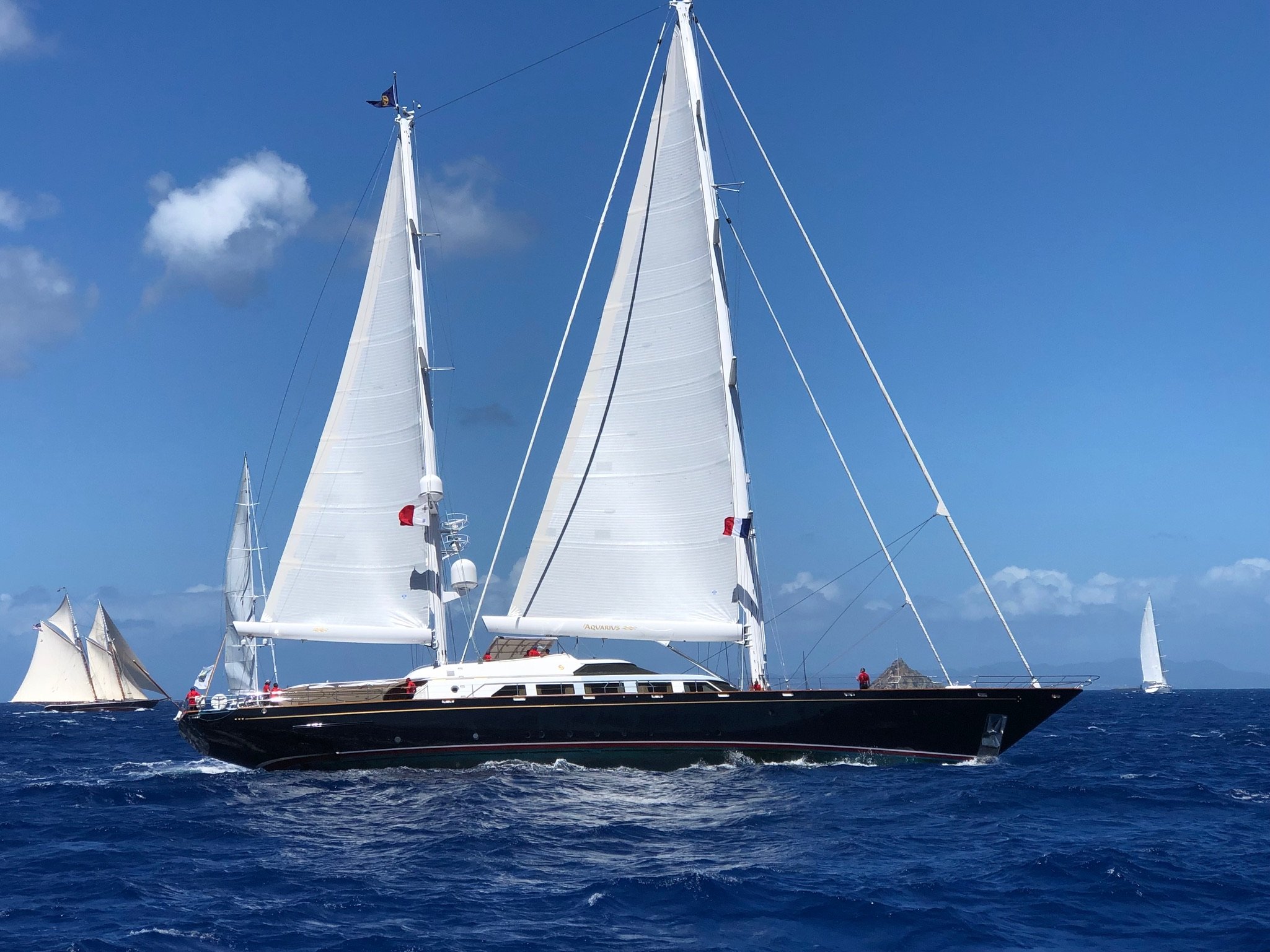 A black sailboat with white sails sailing on the ocean with two smaller sailboats in the distance and a partly cloudy sky.