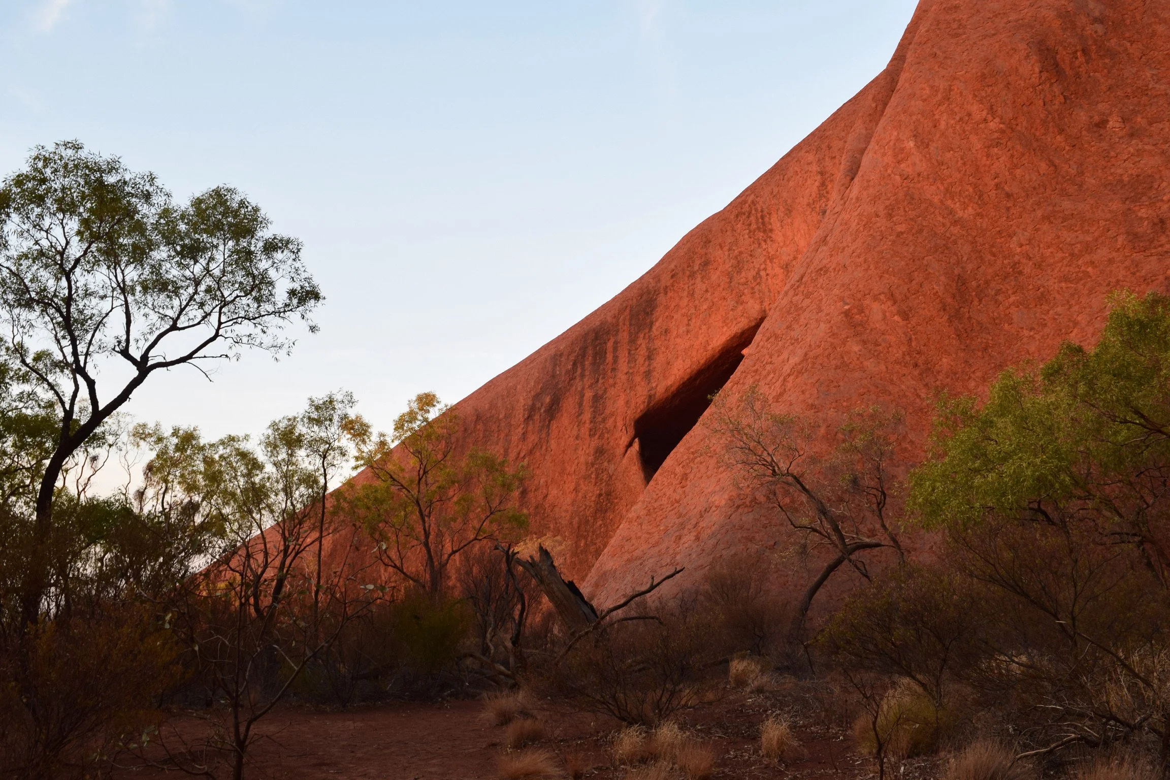 A large red rock formation in a desert landscape with sparse green trees and bushes, under a clear sky.