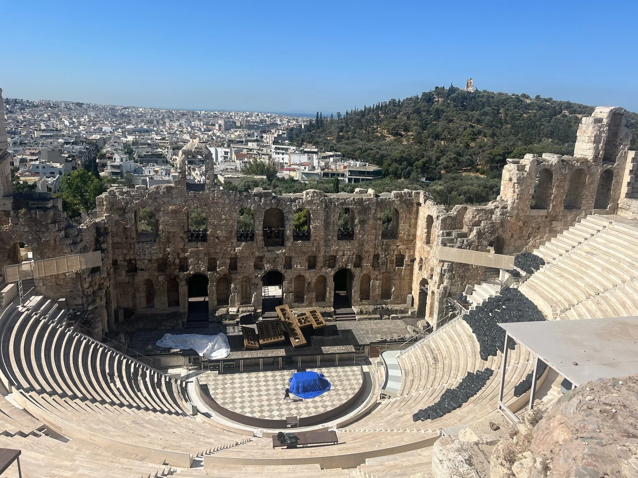 The ancient theater at the Acropolis in Athens, Greece, with ruins and seating, overlooking a cityscape and a hill with a statue.