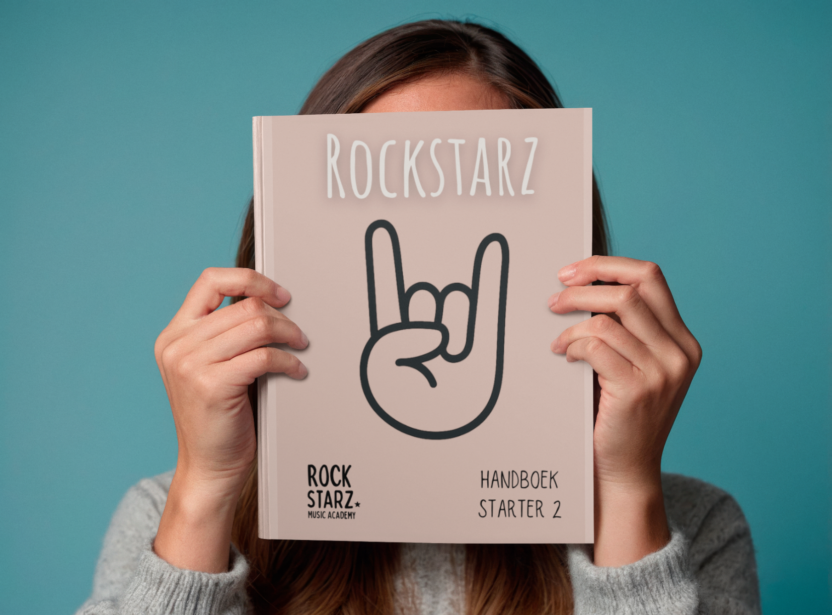Person holding a music book titled 'Rockstarz' in front of their face. The book features a hand making the 'rock on' gesture. The background is teal, and the person is wearing a gray sweater.