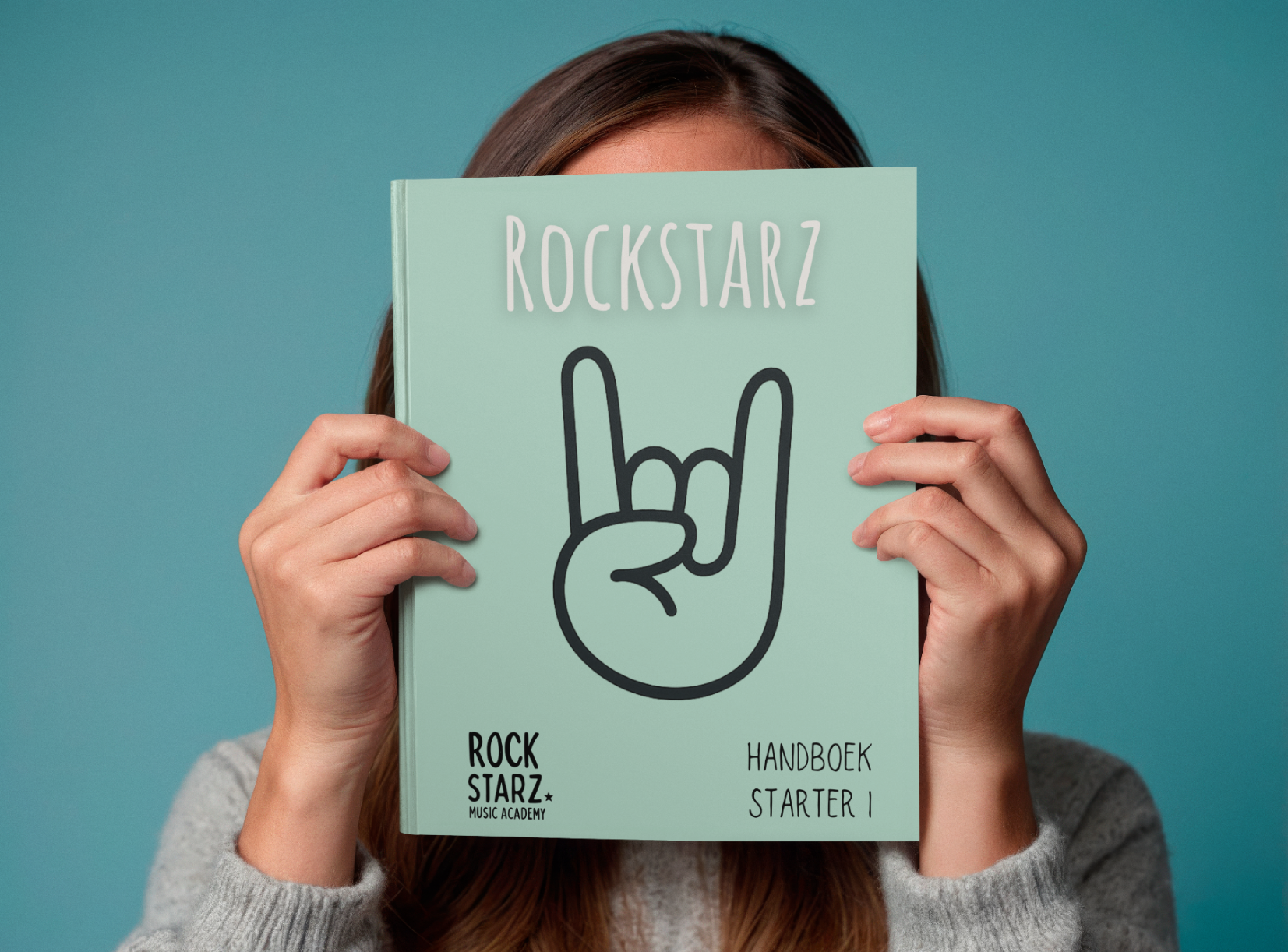 Person holding a booklet titled 'Rockstar' with a rock hand symbol on the cover. The cover includes the text 'Rockstar European Music Academy,' 'Handboek Starter 1.'