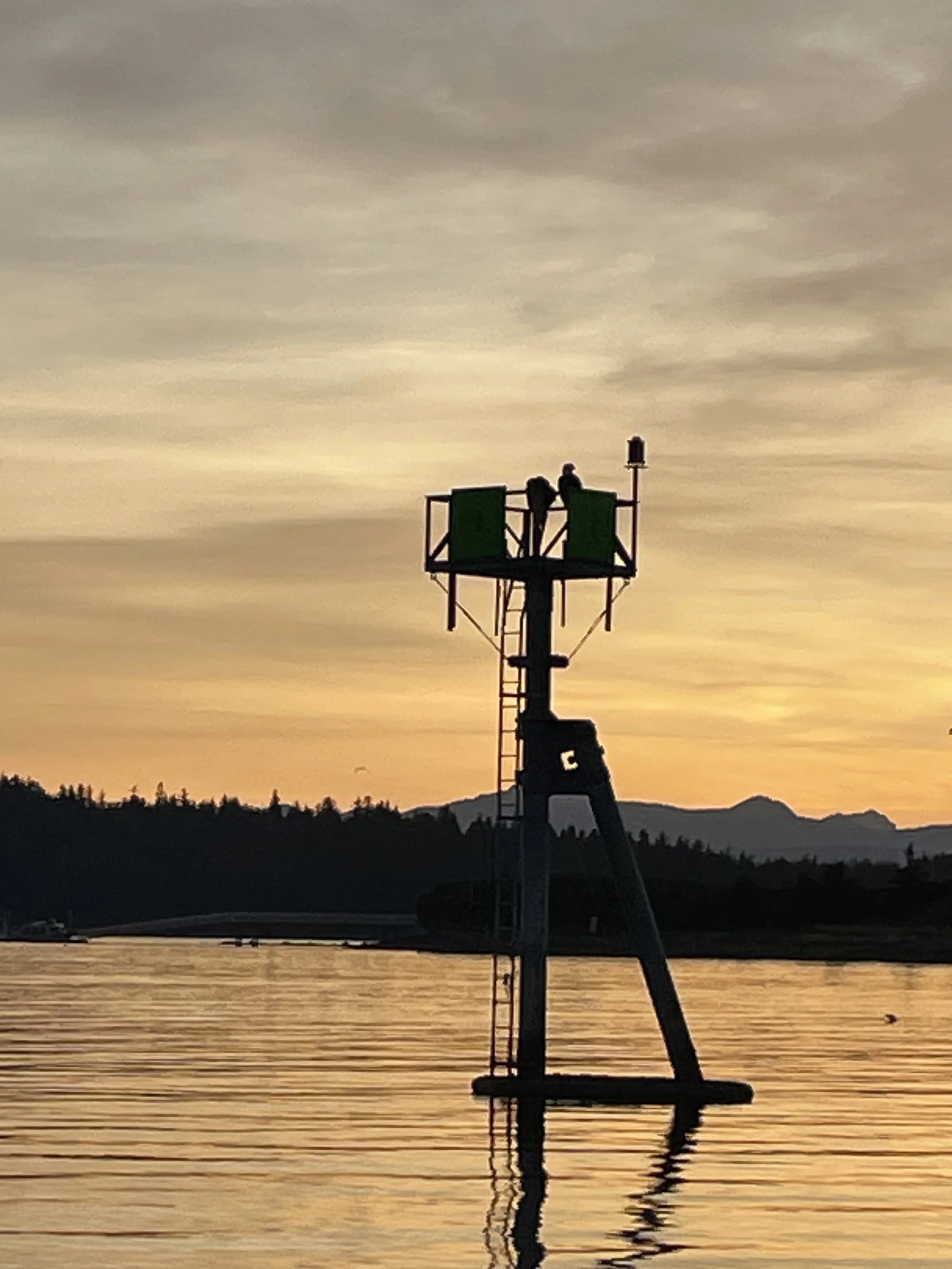 A silhouette of a sculpture on a platform standing in water at sunset, with two figures sitting on the platform and mountains in the background.