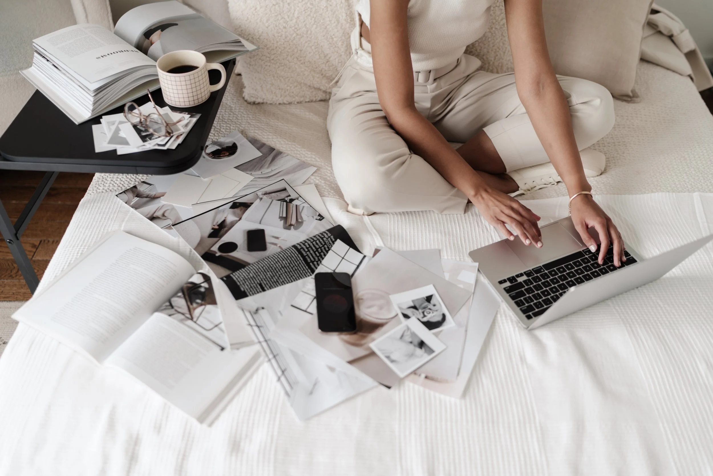 A person sitting cross-legged on a bed, working on a laptop. The bed is cluttered with open books, photographs, magazines, glasses, a cup of coffee, and some electronic devices. There is a small table next to the bed with more books, glasses, and a cup of coffee.