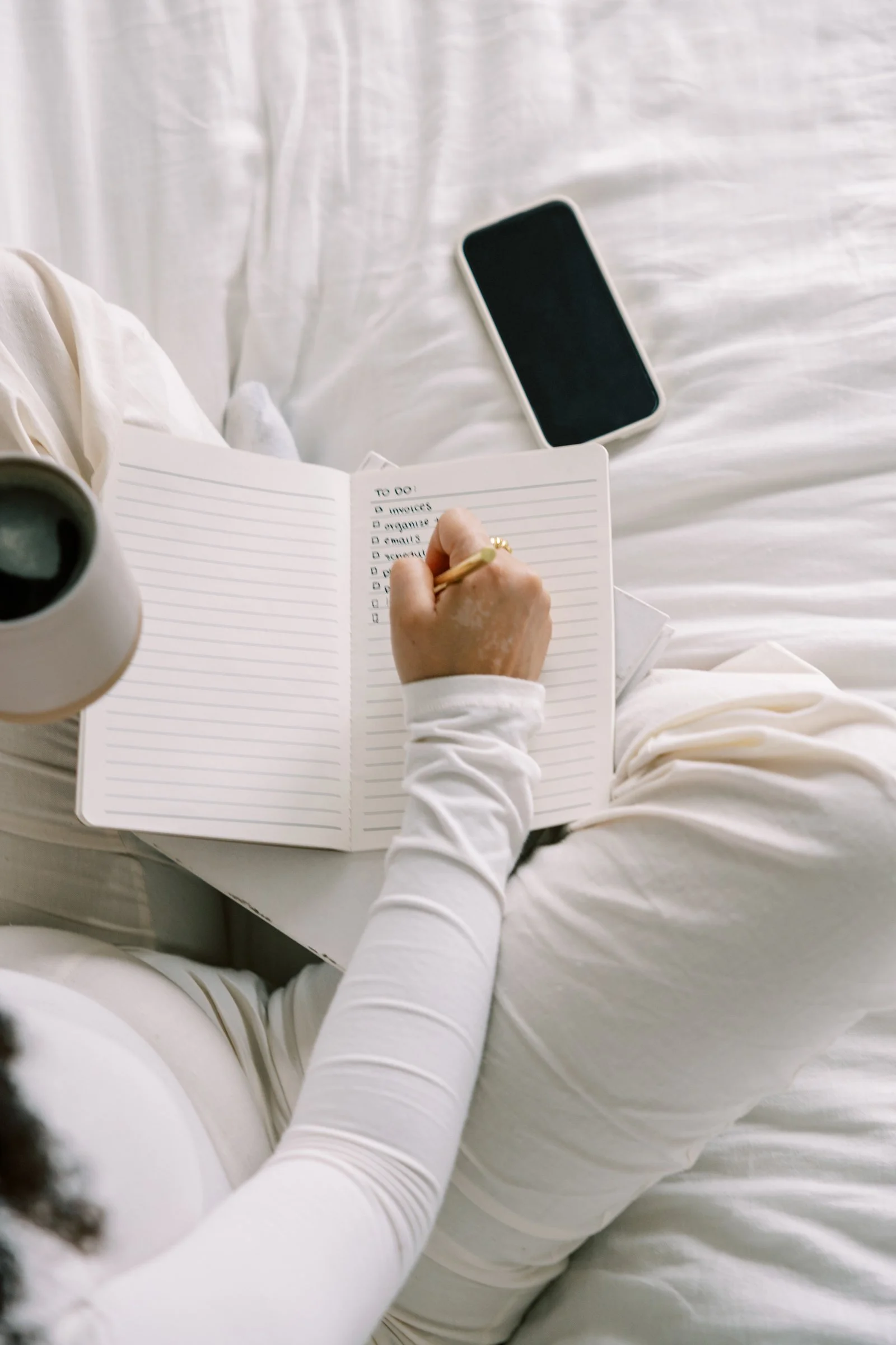 Person sitting on a bed, writing in a notebook with a list of tasks, with a smartphone and a coffee mug nearby.