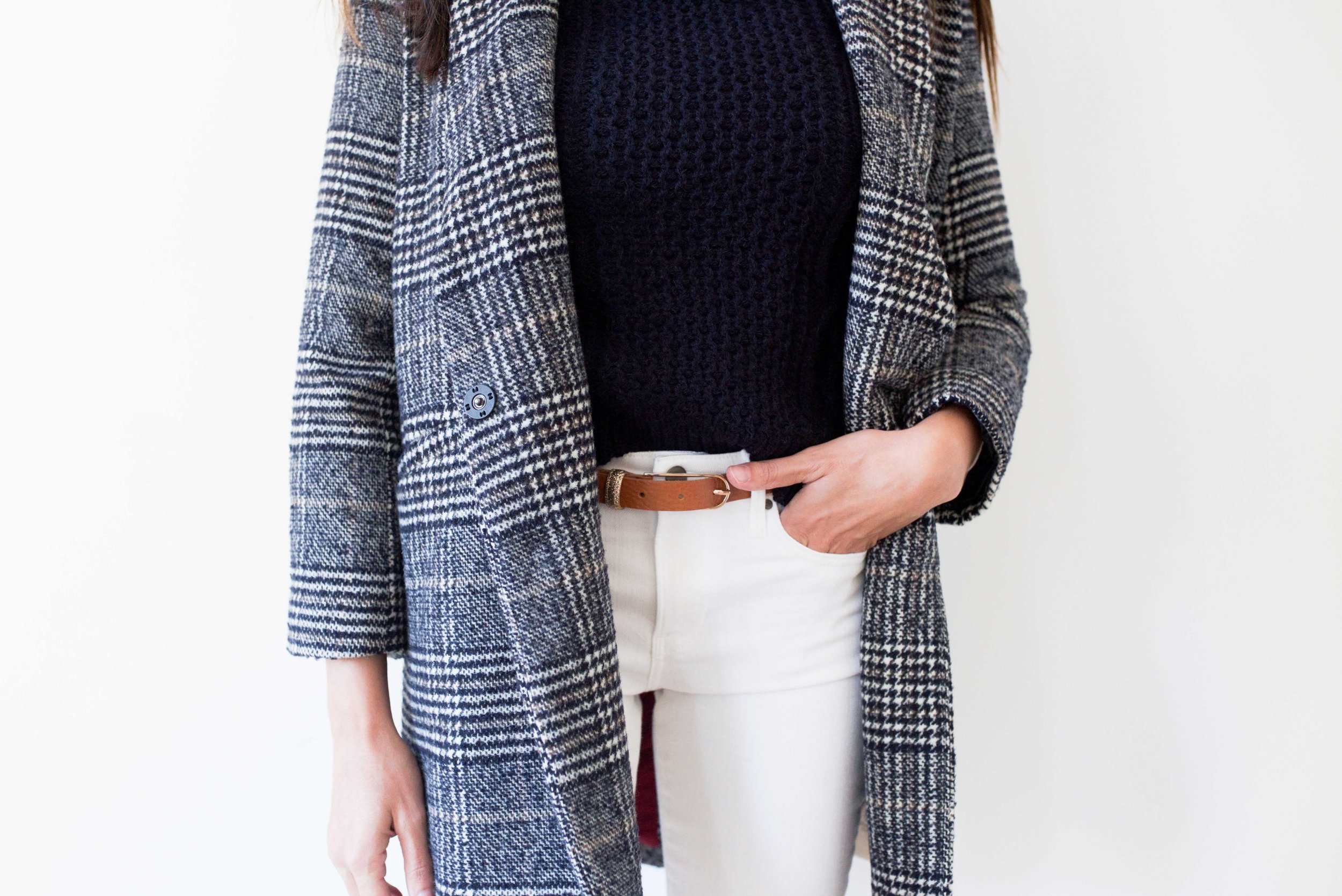 Close-up of a woman wearing a black knit sweater, a gray and black houndstooth patterned coat, and white pants with a brown belt.