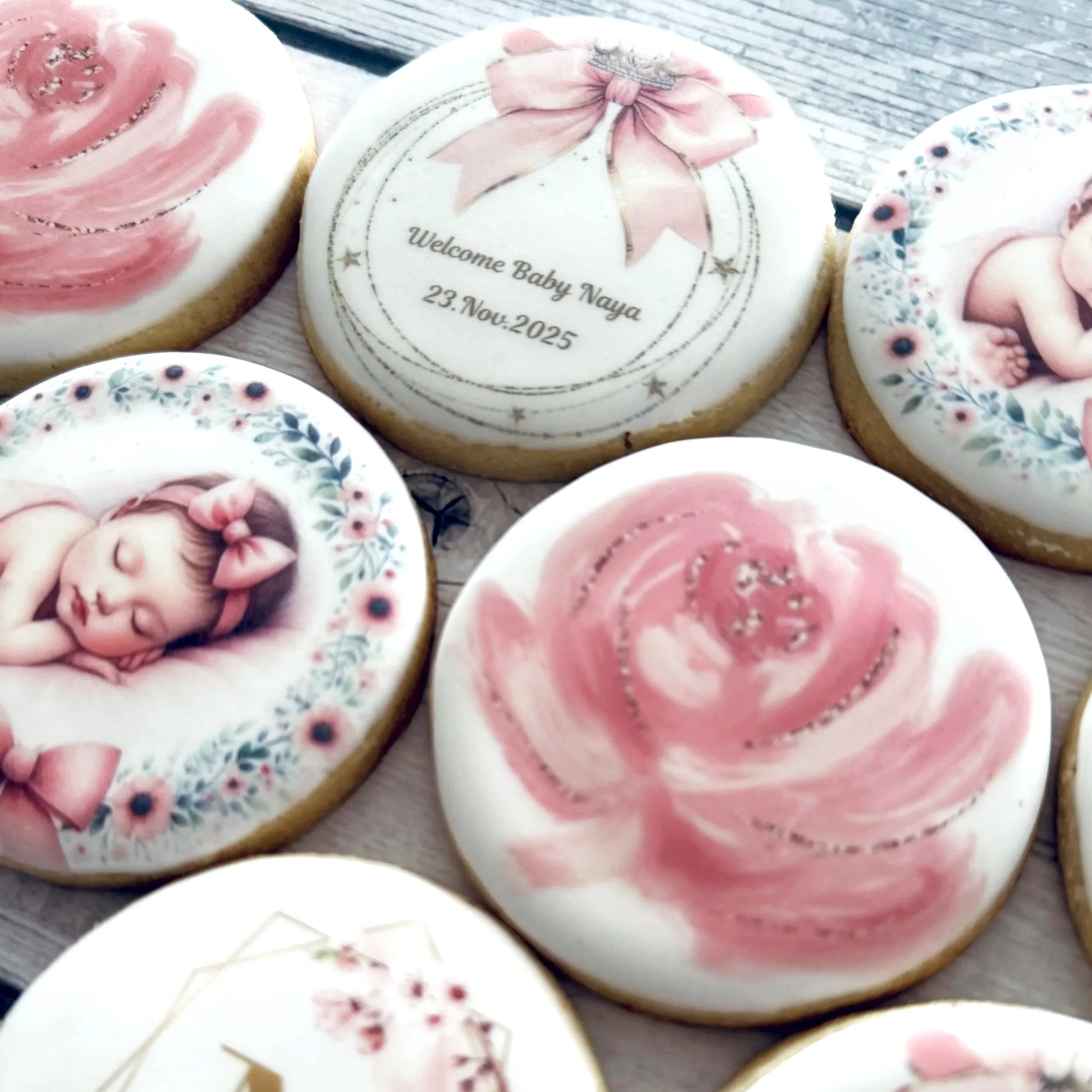 Decorative cookies with baby-themed designs and messages, featuring portraits of a sleeping baby girl with a pink bow, floral wreaths, and a message welcoming baby Naya on November 23, 2025.
