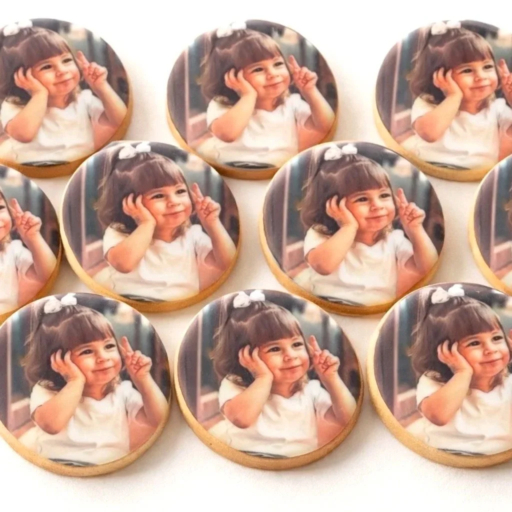 A gloved hand holding a round button with a picture of a young girl with dark hair and bangs, wearing a white shirt, making a playful gesture with her fingers near her face. Multiple similar buttons with the same picture are stacked in the background, all sealed in plastic packaging.