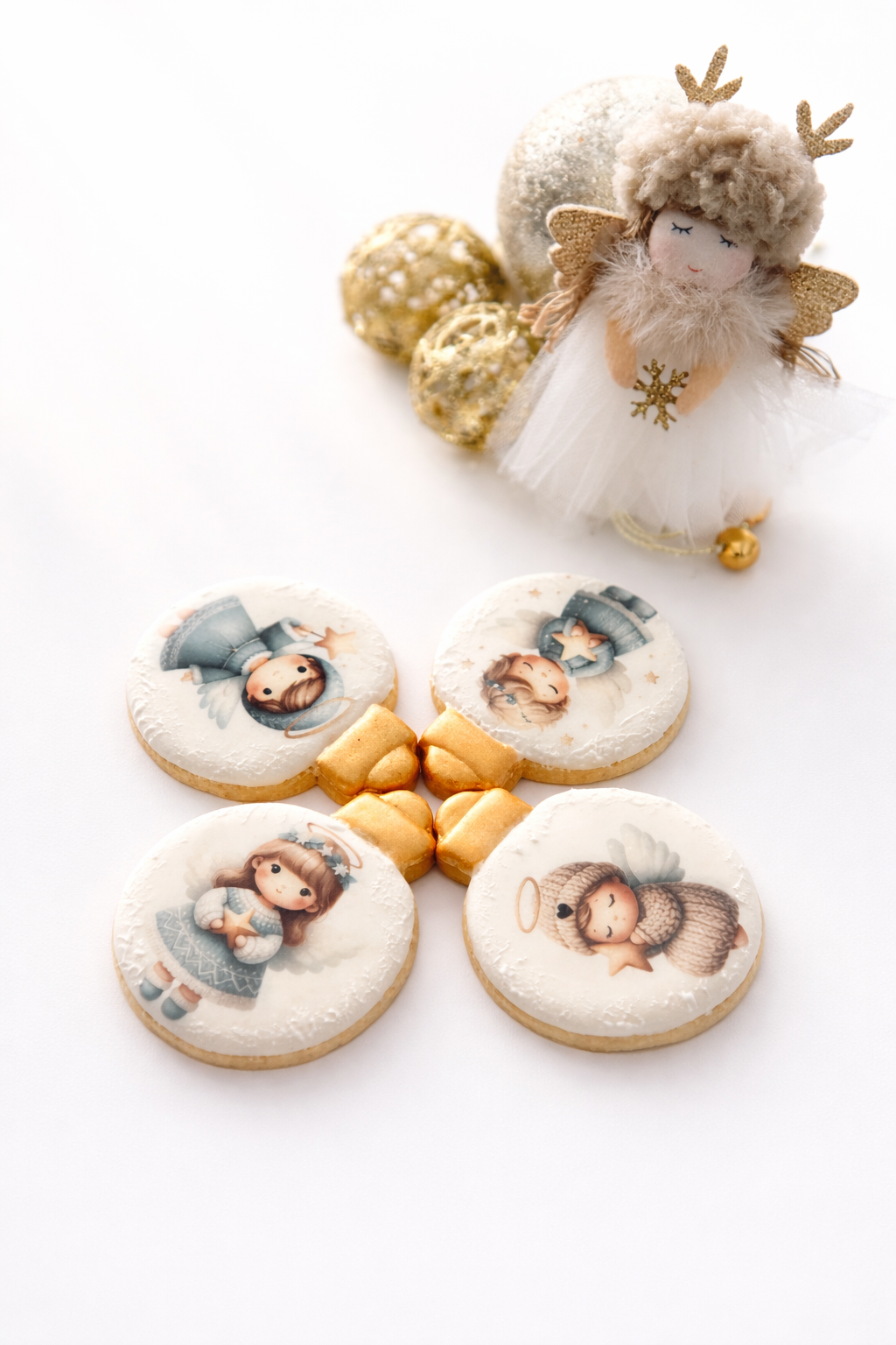 Christmas-themed decorations including a plush angel doll with a fluffy gray hat, glittery antlers, golden wings, and a white tutu, along with four cookies decorated with cute angel illustrations and golden ribbon-shaped cookies, on a white wooden surface.