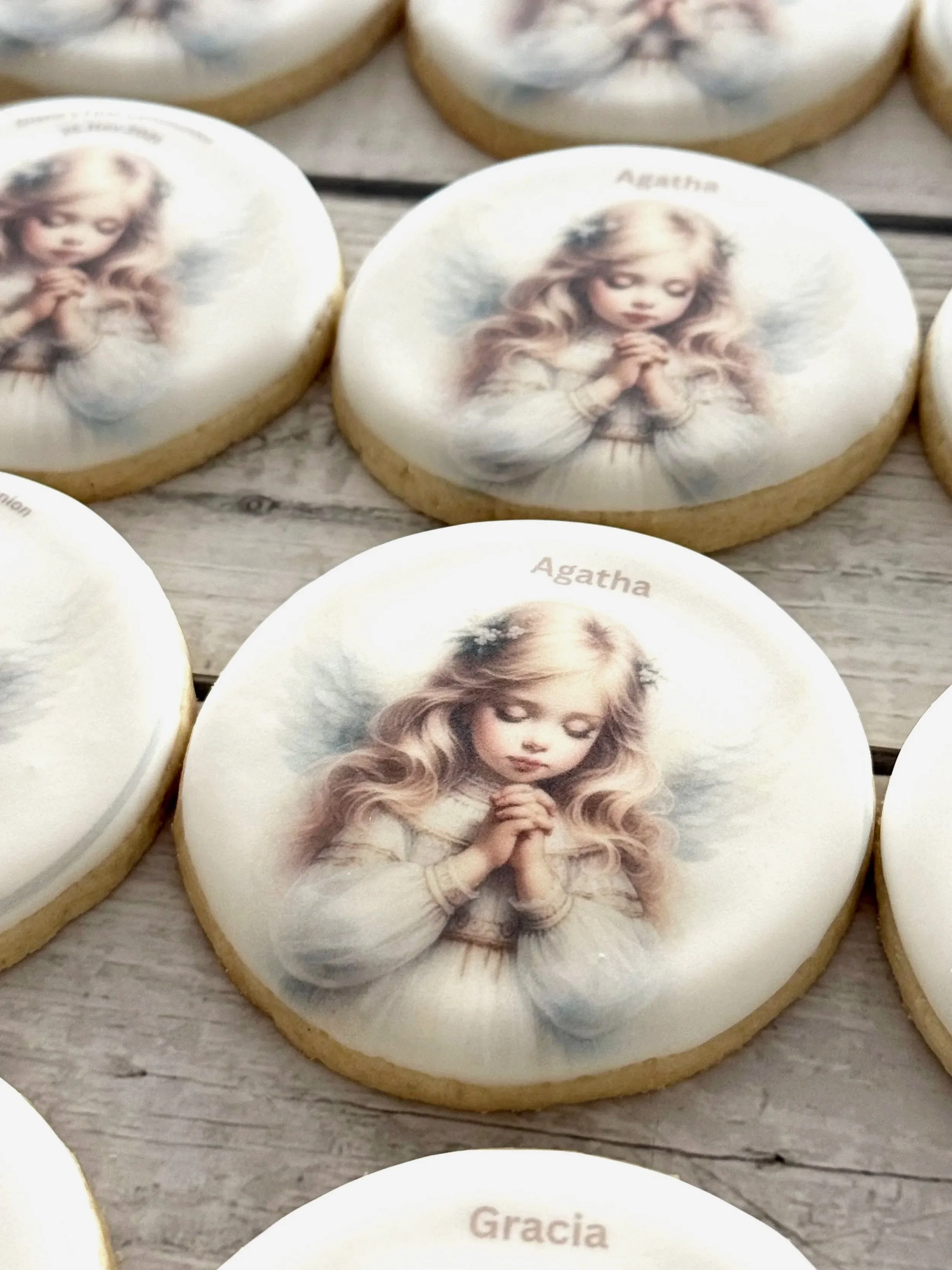 Cookies decorated with an image of a young girl with long blonde hair, praying with hands clasped, on a white background, with names "Agatha" and "Gracia" inscribed on some cookies.