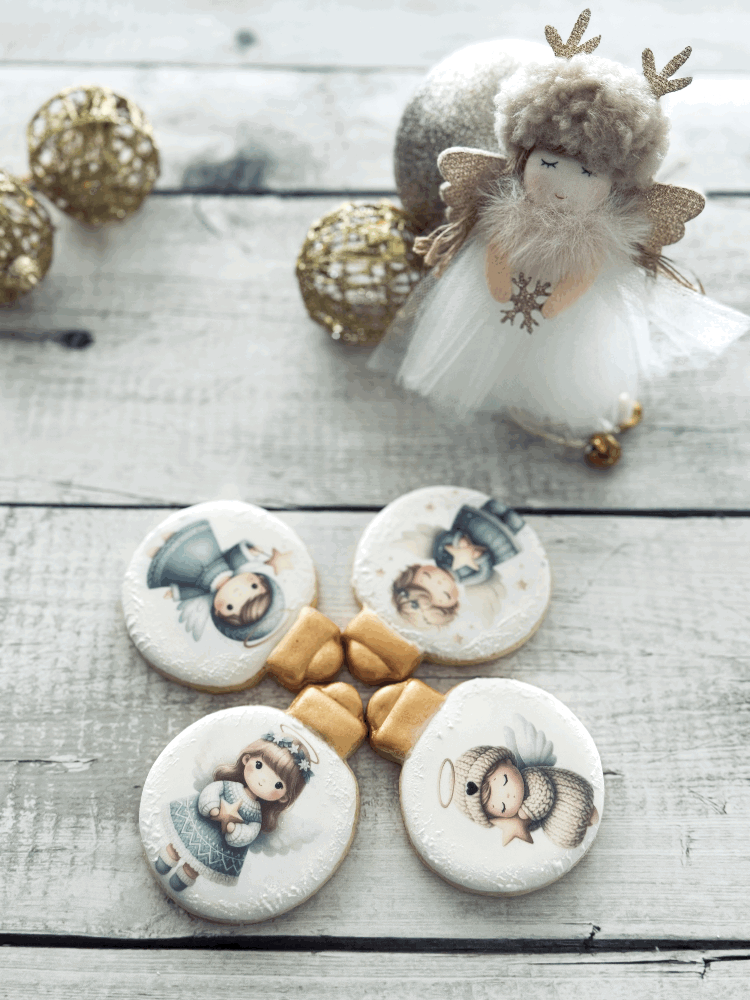 Christmas-themed decorations including a plush angel doll with a fluffy gray hat, glittery antlers, golden wings, and a white tutu, along with four cookies decorated with cute angel illustrations and golden ribbon-shaped cookies, on a white wooden surface.
