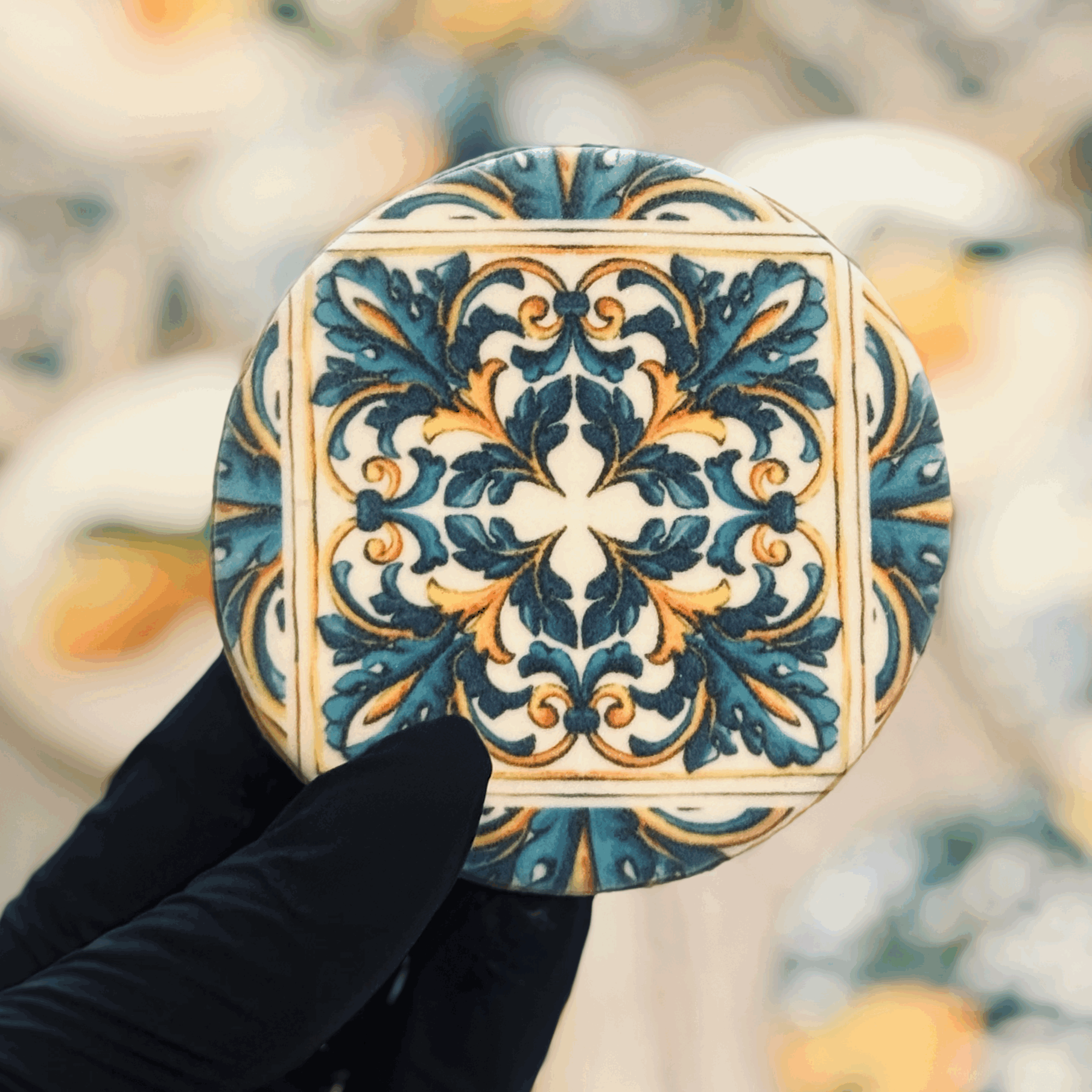A person wearing a black glove holding a decorative ceramic tile with a symmetrical blue, yellow, and white floral pattern.