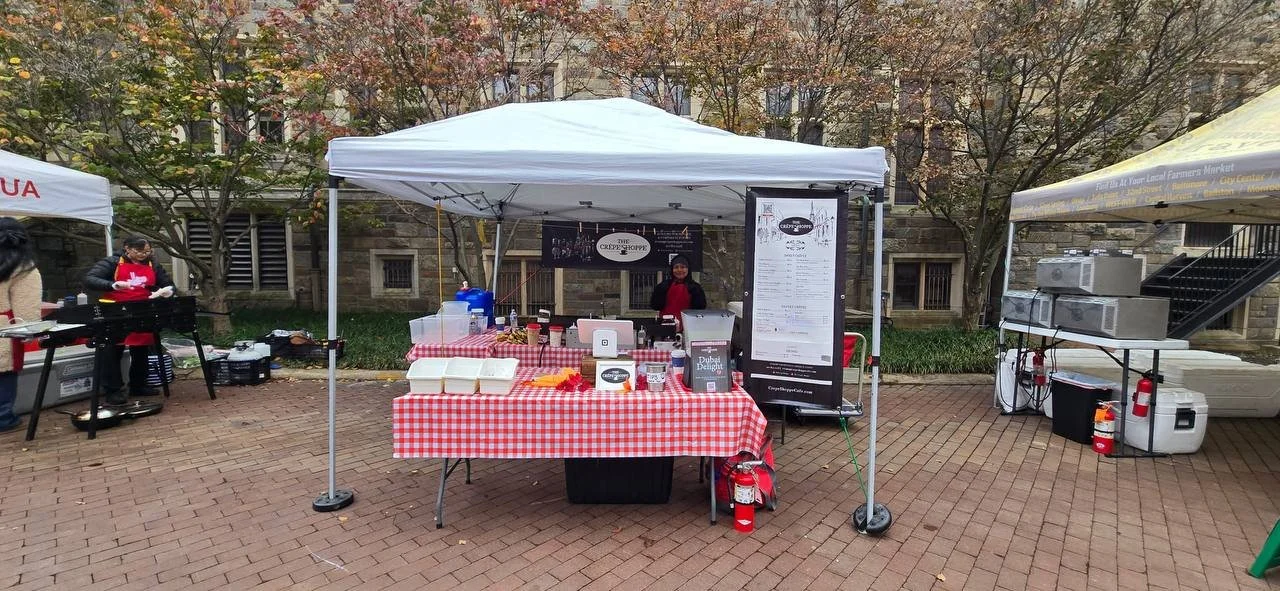 A food vendor stall at an outdoor market set up with a white canopy tent, red and white checkered tablecloth, and various food containers and condiments. Two women are working at the stall, and a table with a grill is nearby on the left. Behind the stall are leafless trees and a brick building.