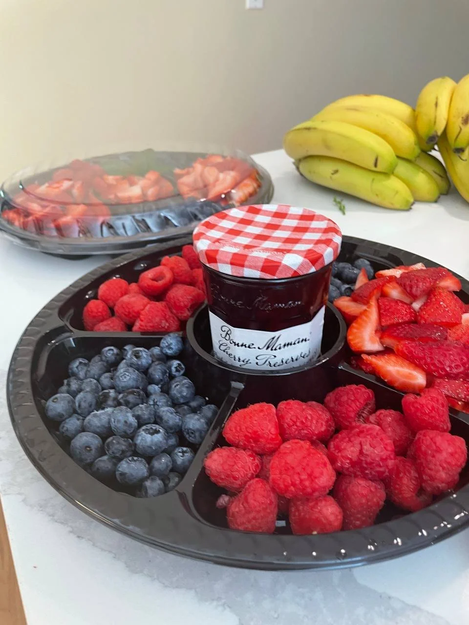 A fruit platter with blueberries, raspberries, strawberries, and a jar of cherry preserves, with bananas and sliced strawberries on the side.