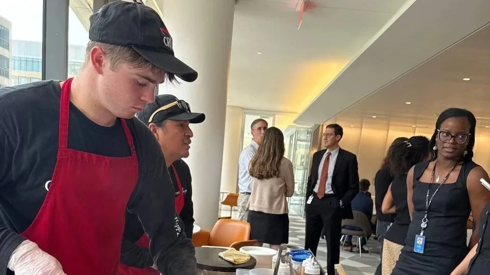 People attending an indoor event, with some preparing or serving food at a table. Two young men are in the foreground, one wearing a red apron and the other in black, focused on their tasks. In the background, several adults are standing and conversing near large windows.