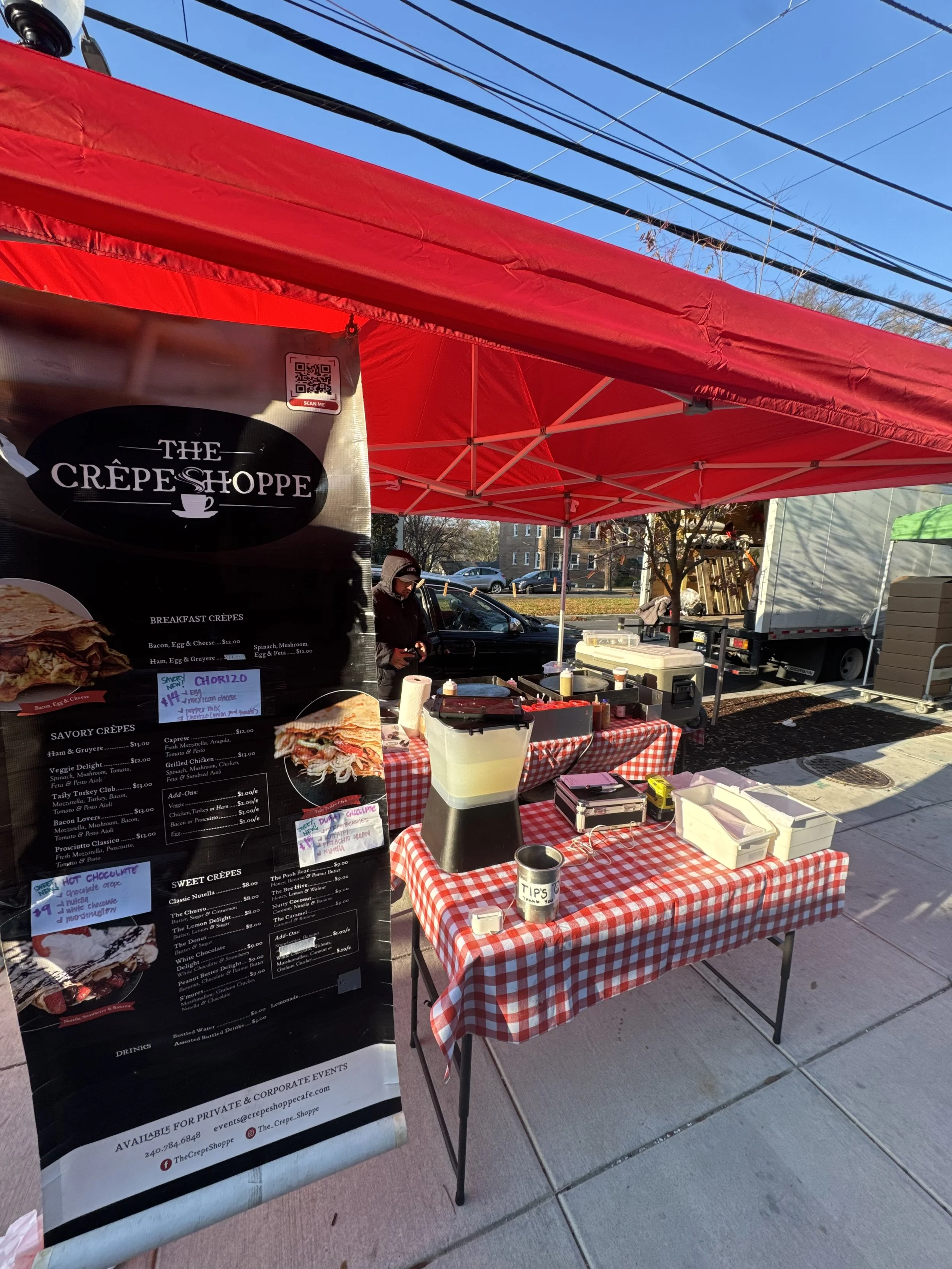 Food stand with a red canopy selling crepes, with a black menu board displaying various crepe options, and a table with toppings and utensils on a checkered tablecloth.