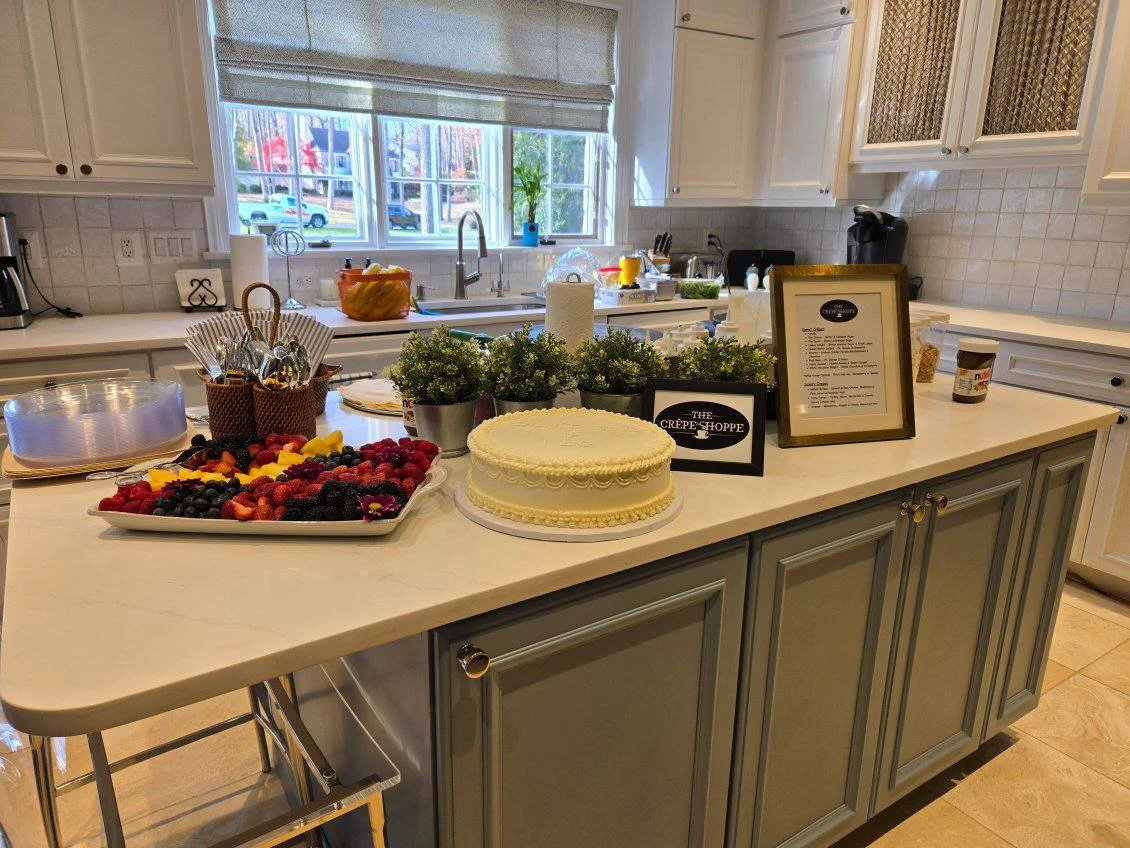 A kitchen island with a cake, a tray of mixed berries and fruit, potted plants, and framed signs. The kitchen has white cabinets and a window above the sink.