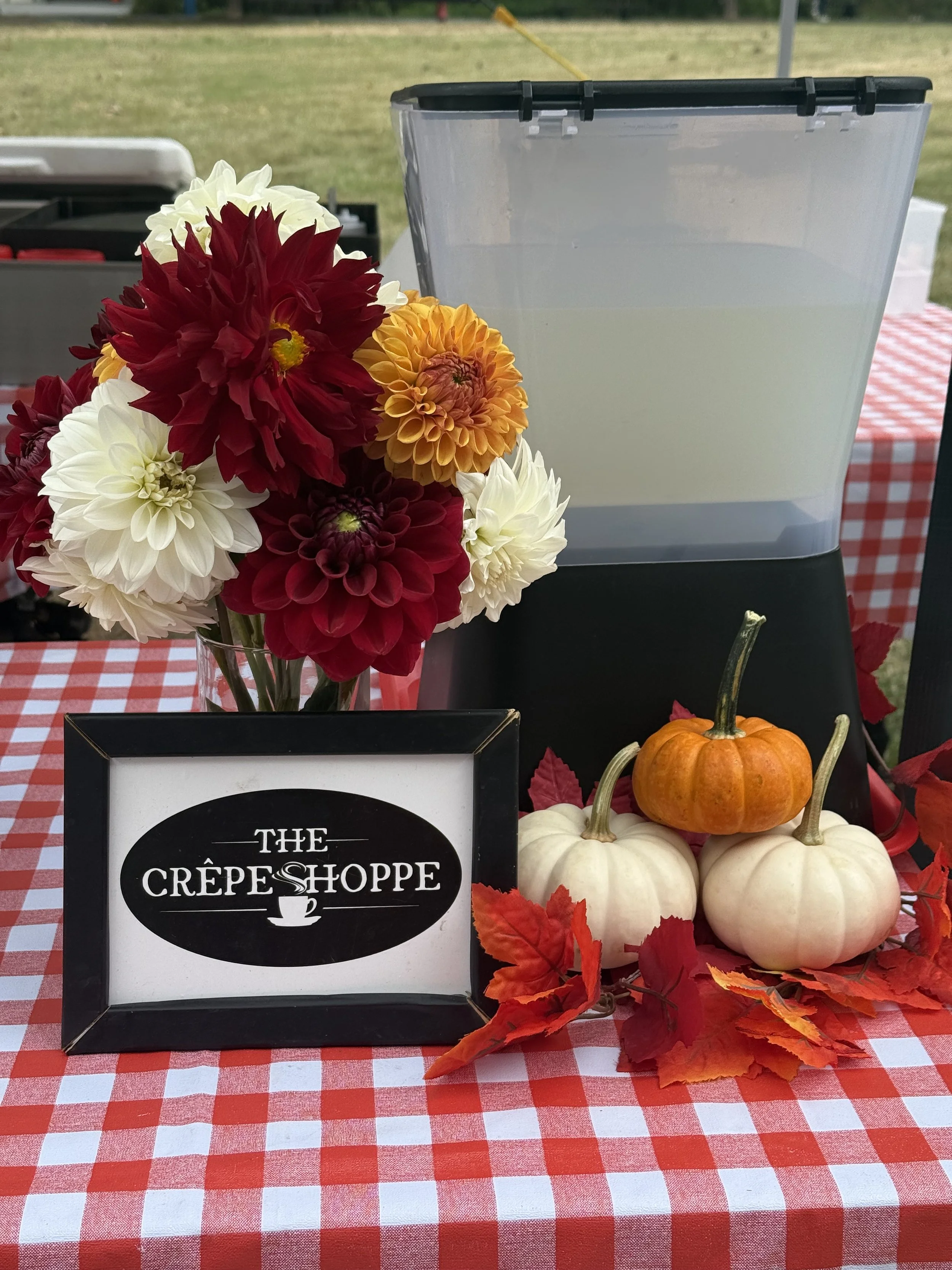 A table with a red and white checkered tablecloth displaying a bouquet of multicolored dahlias, a small black-framed sign that reads 'The Crêpe Shoppe,' three small pumpkins (two white and one orange), and some red autumn leaves.