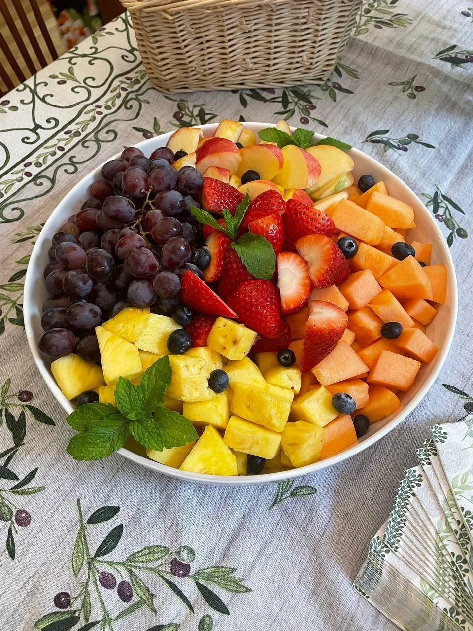 Assorted fresh cut fruits in a white bowl, including grapes, strawberries, pineapple, cantaloupe, honeydew, blueberries, and apple slices, garnished with mint leaves.