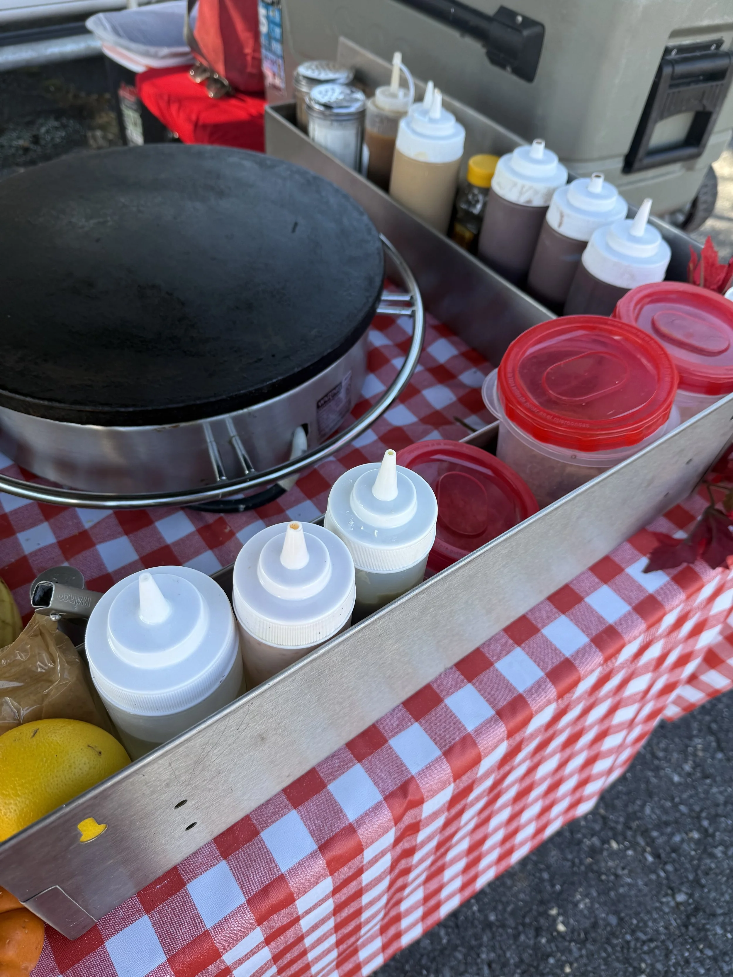 A street food cart with a black cooking surface, condiment bottles, and containers with lids, set on a red and white checkered tablecloth.