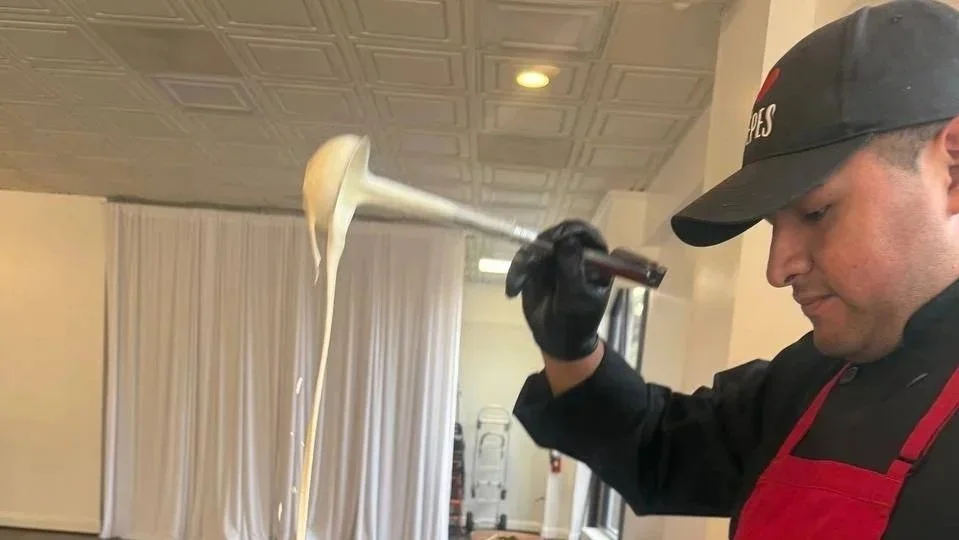 Chef at The Crêpe Shoppe Café wearing a black shirt, red apron, and black gloves, lifting a spoon with rich melted cheese dripping down while preparing a fresh savory crêpe for a private catering birthday party in Washington DC