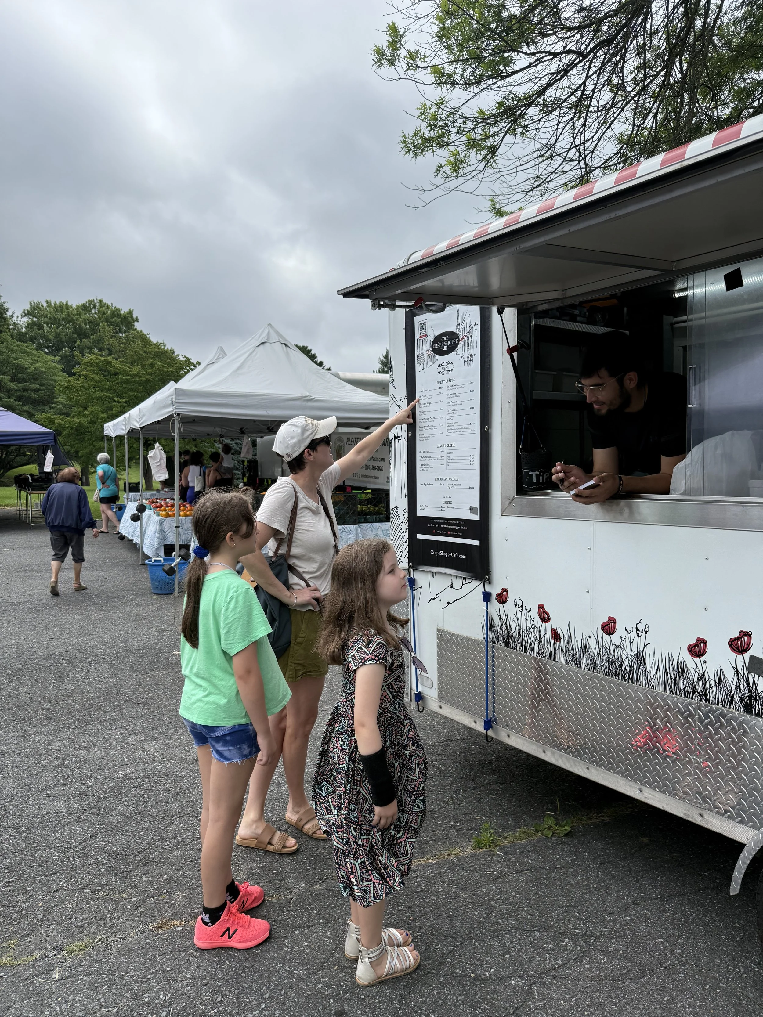 People placing orders at a food truck with a menu, at an outdoor market with tents and vendors in the background.